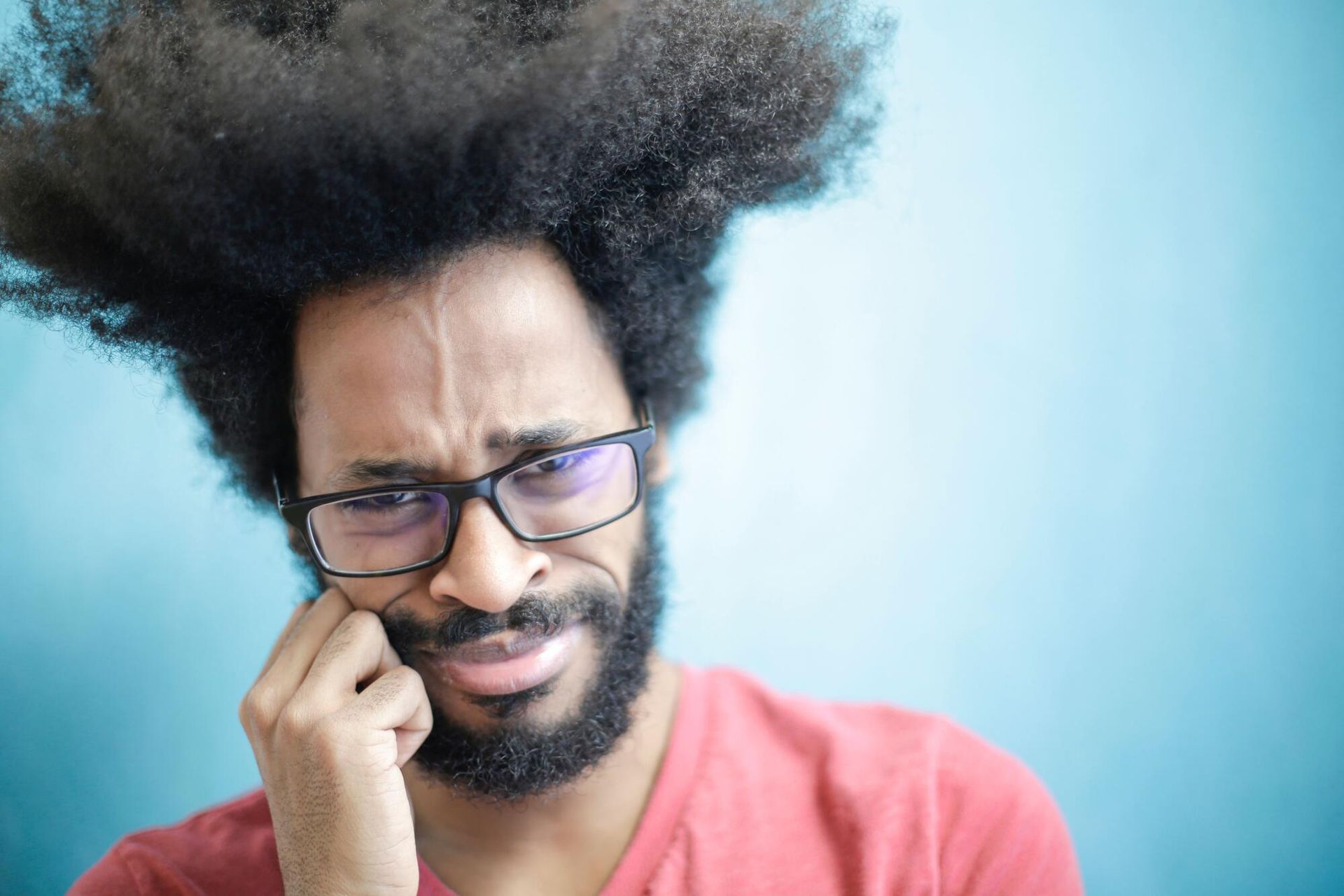 Man with glasses grimacing and scratching his cheek against a blue background