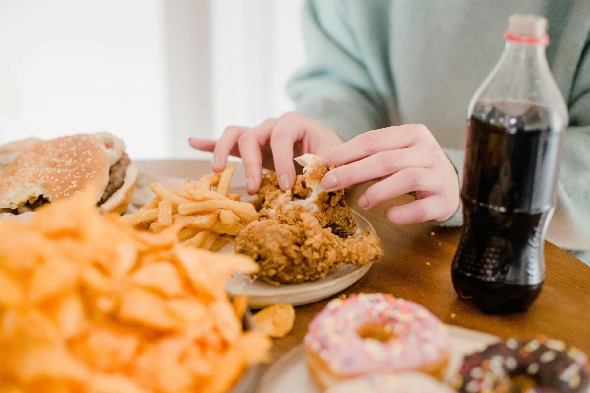Hands eating fast food with fries, burgers, donuts, and a soda bottle on a table