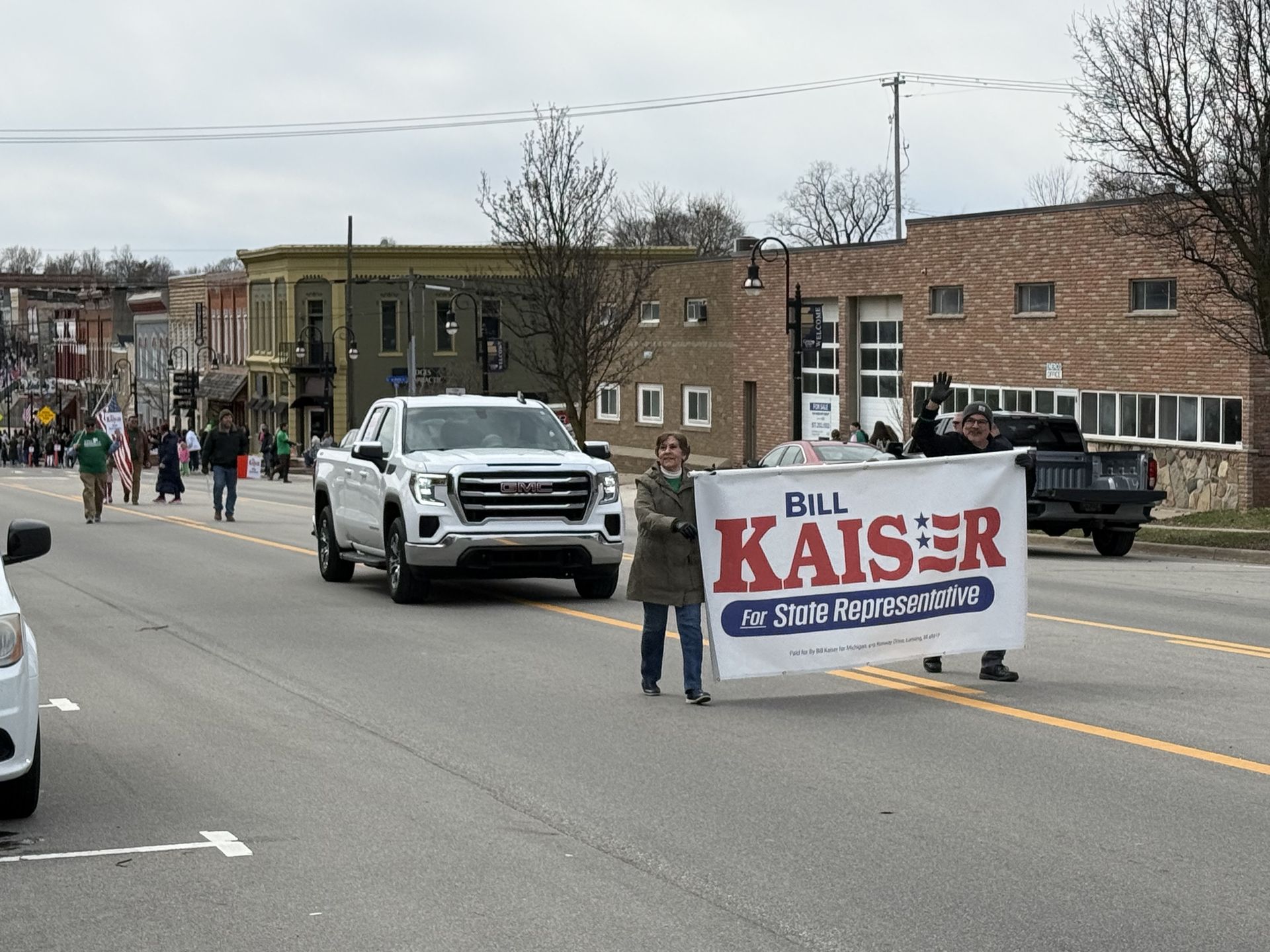 Parade for st. Patrick's day Bill Kaiser banner on a city street beside a white pickup truck.