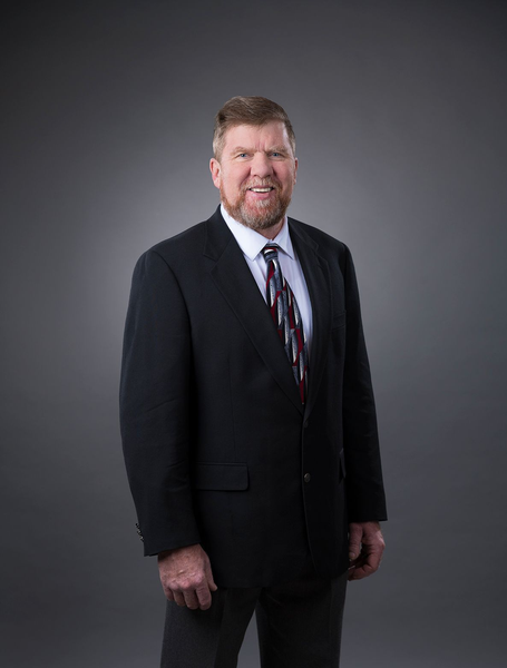 Man in a black suit with a patterned tie, smiling against a gray backdrop.