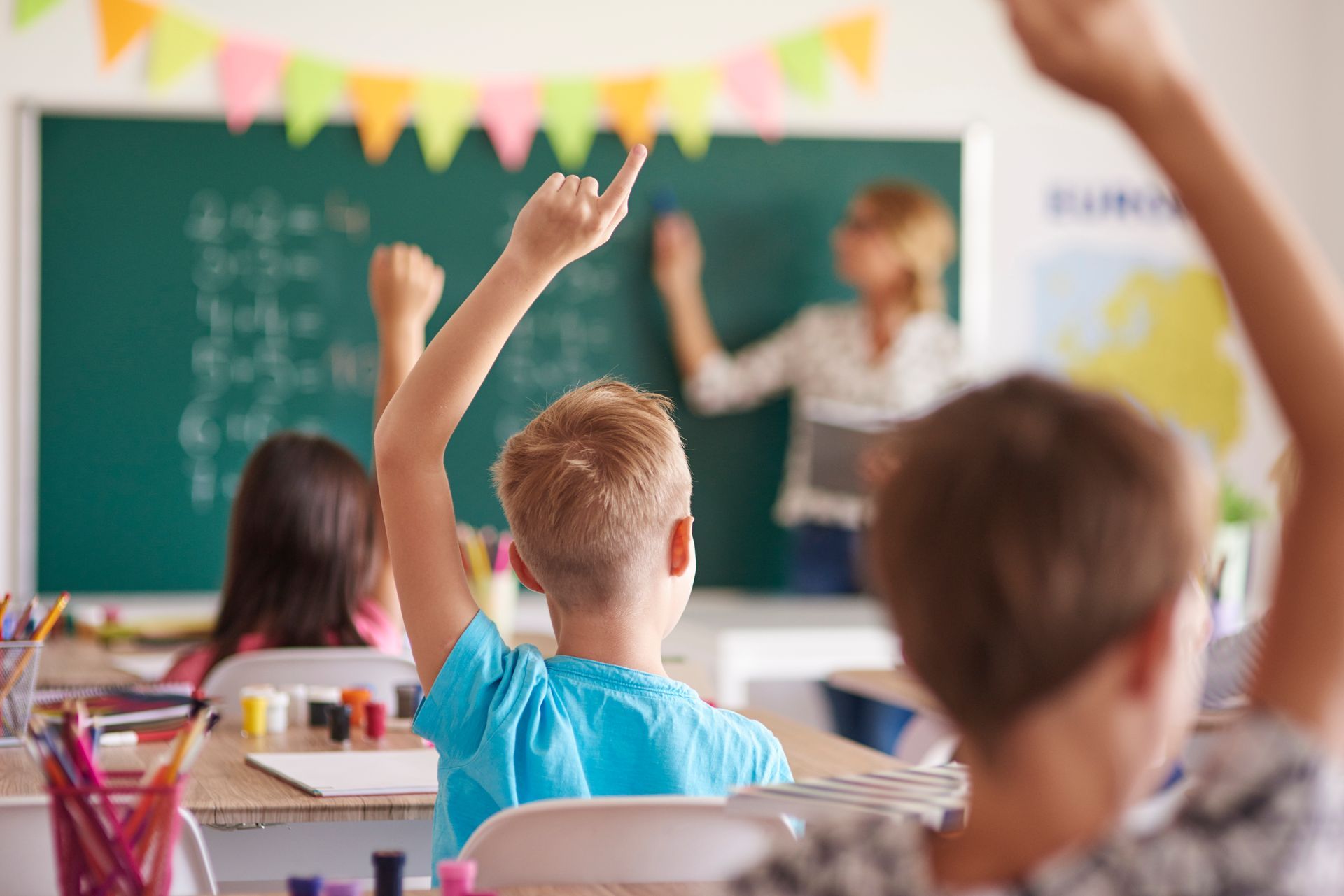 Students raising hands in a classroom, teacher at chalkboard.