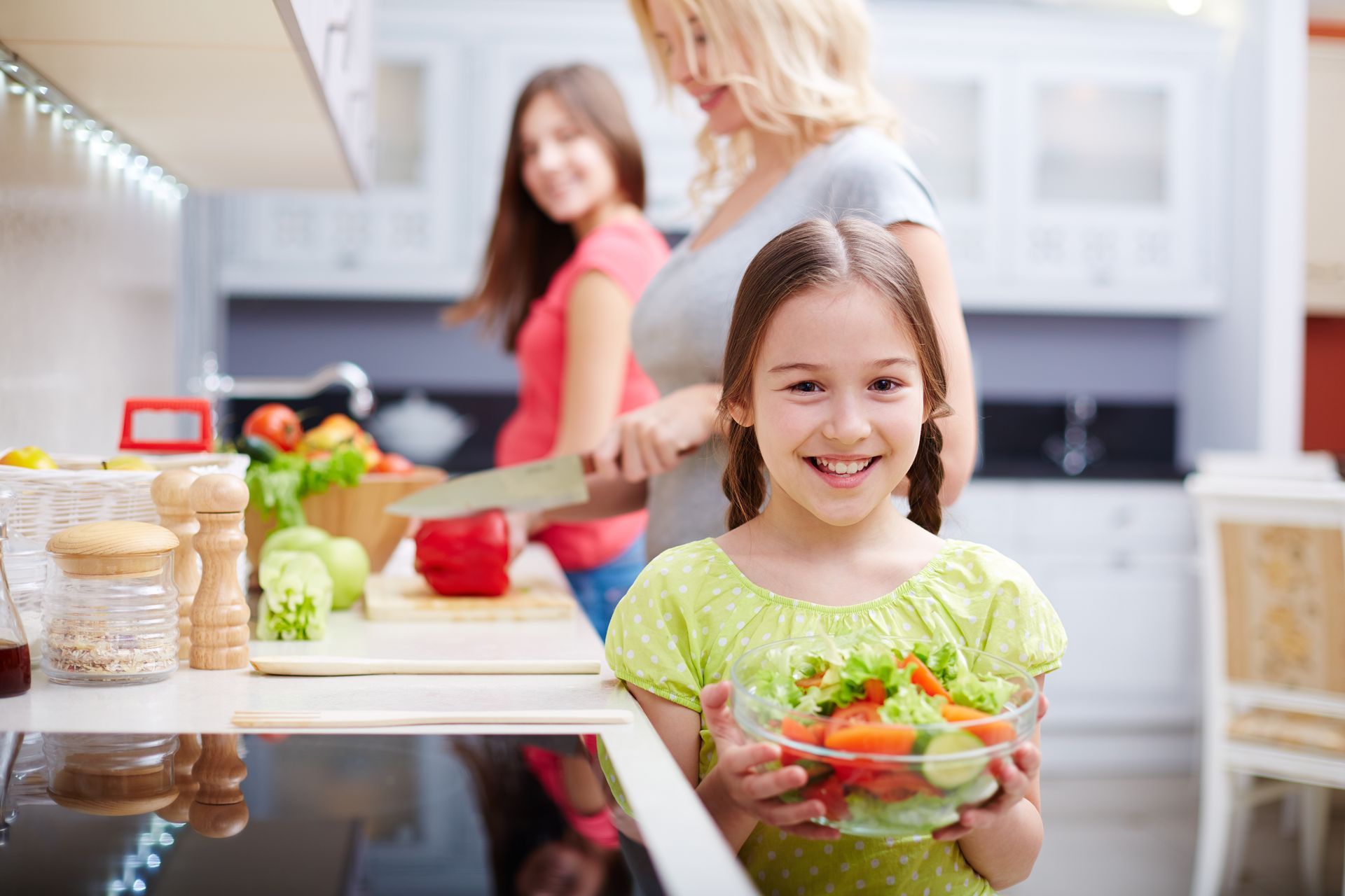 A little girl is holding a bowl of salad in a kitchen.