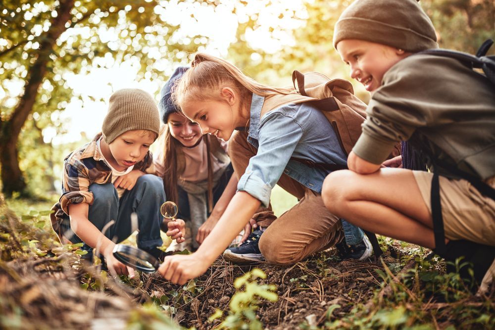Happy children boys and girls making bonfire with magnifying glass together during school camping activity on sunny day