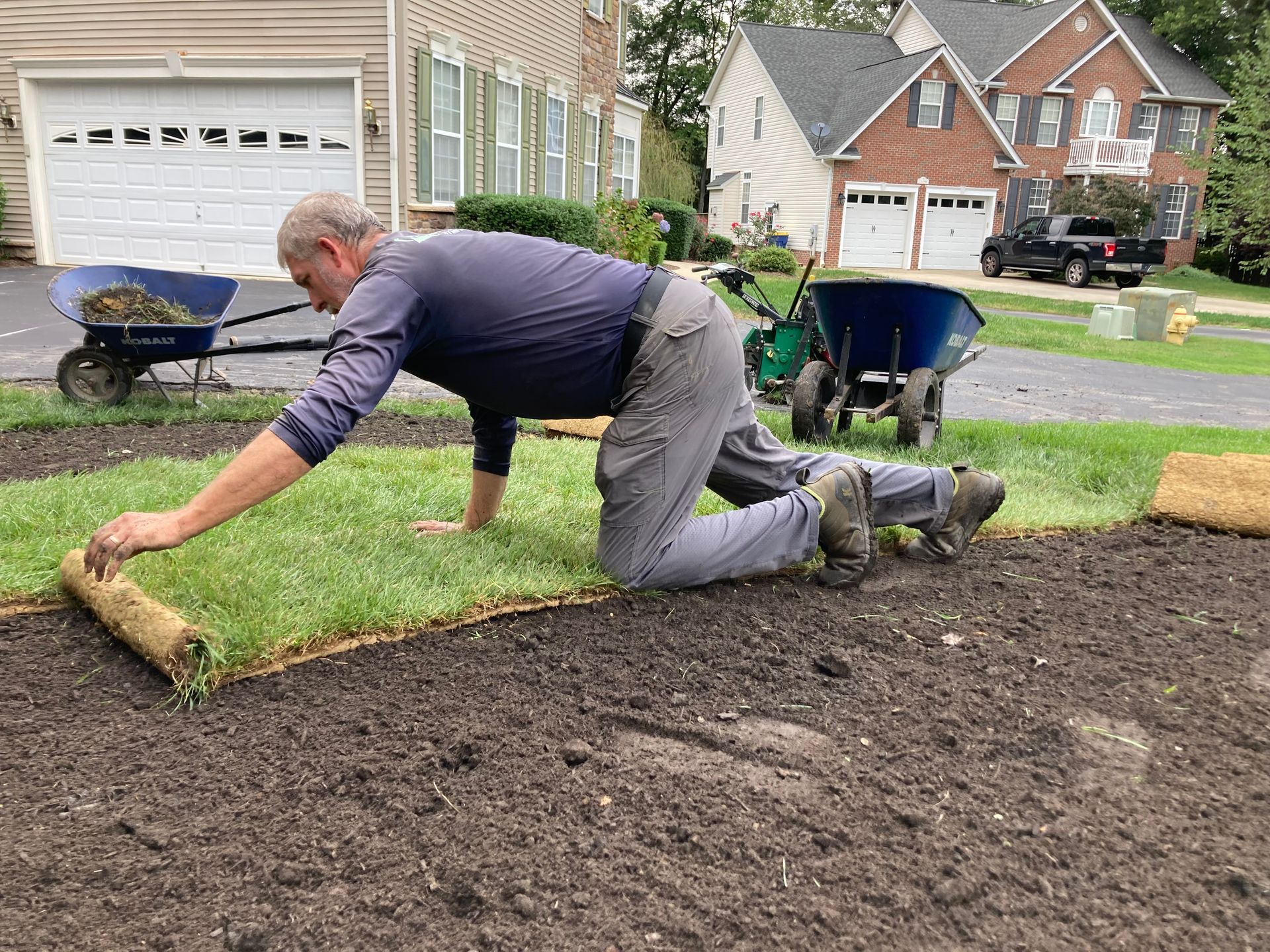 Man laying sod in a front yard beside a house, with a wheelbarrow and fresh soil nearby.