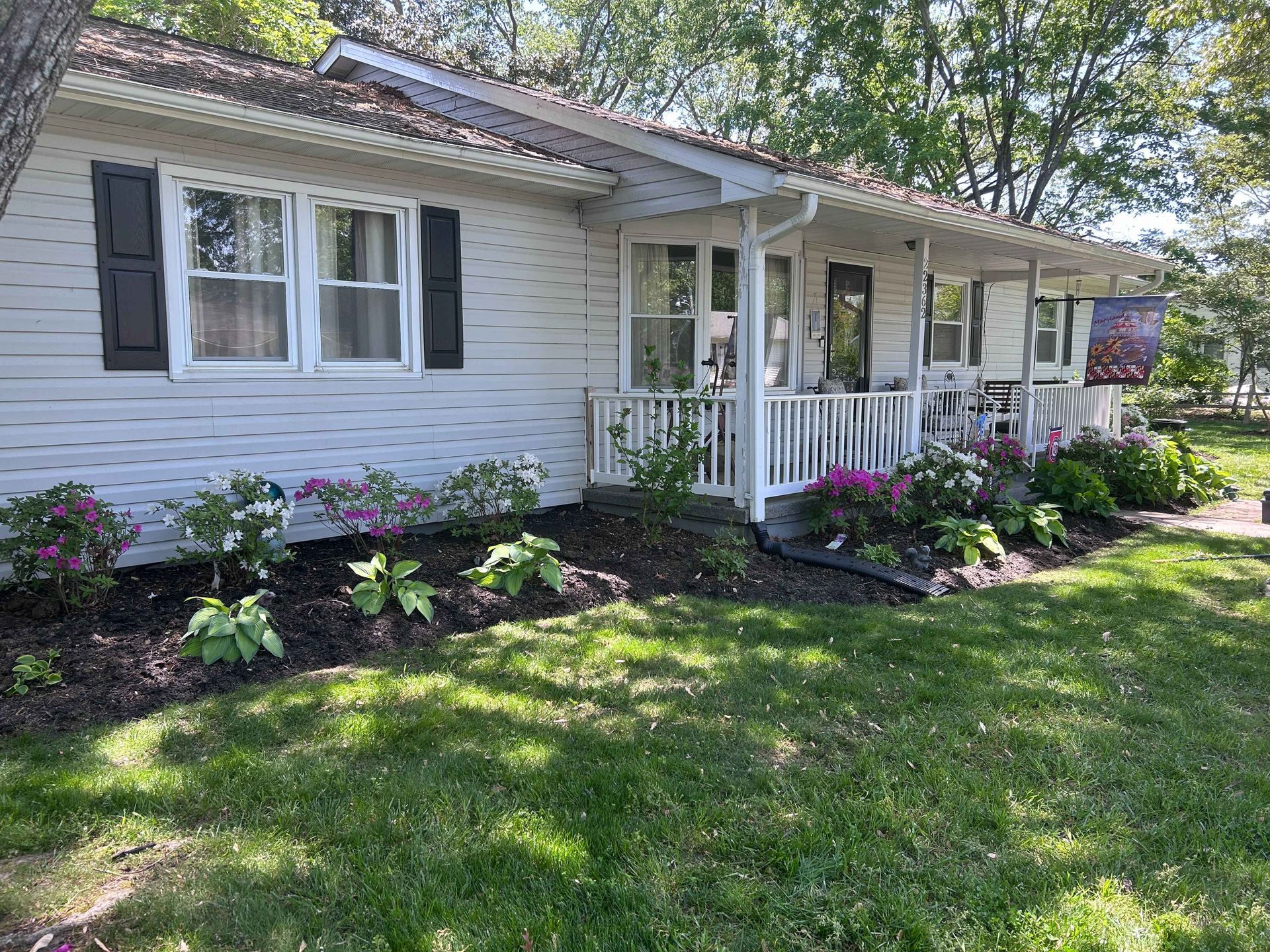 Single-story white house with front porch, flower beds, and a green lawn under trees
