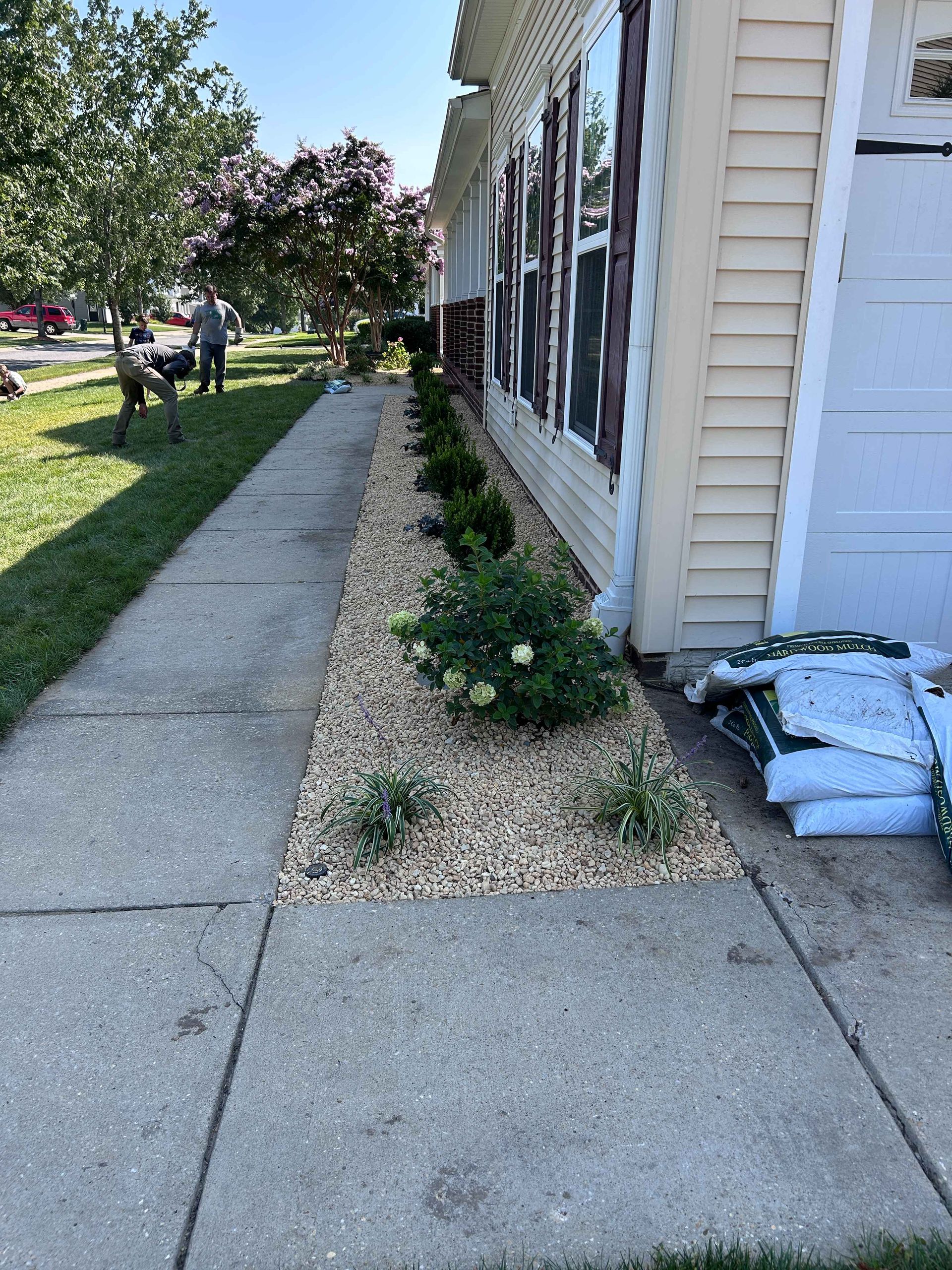 Sidewalk beside a house with gravel beds, shrubs, and stacked bags near the wall; people in the distance.