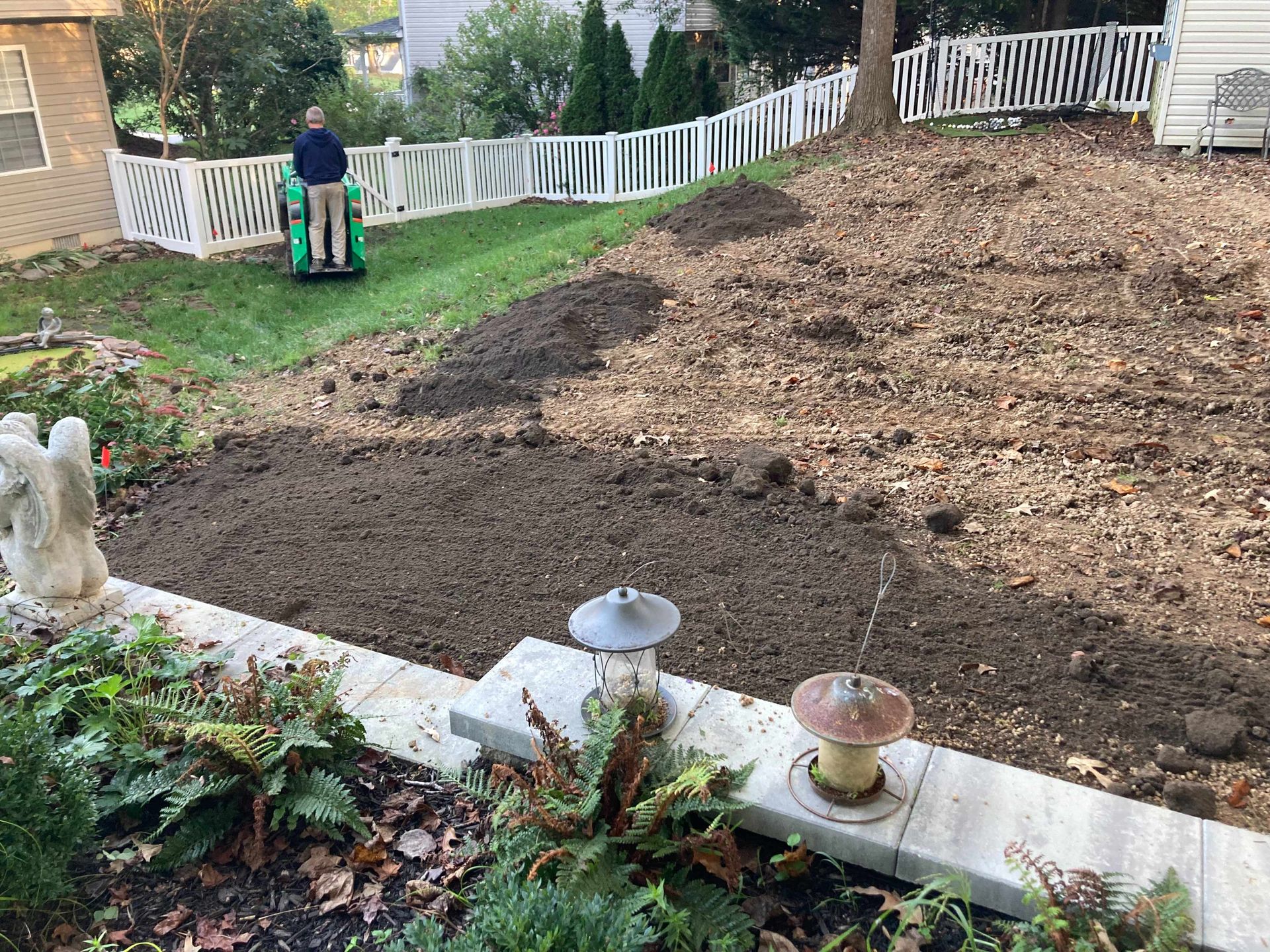 Backyard with fresh dark soil, mulch-covered slope, and a child on a green toy near a white fence