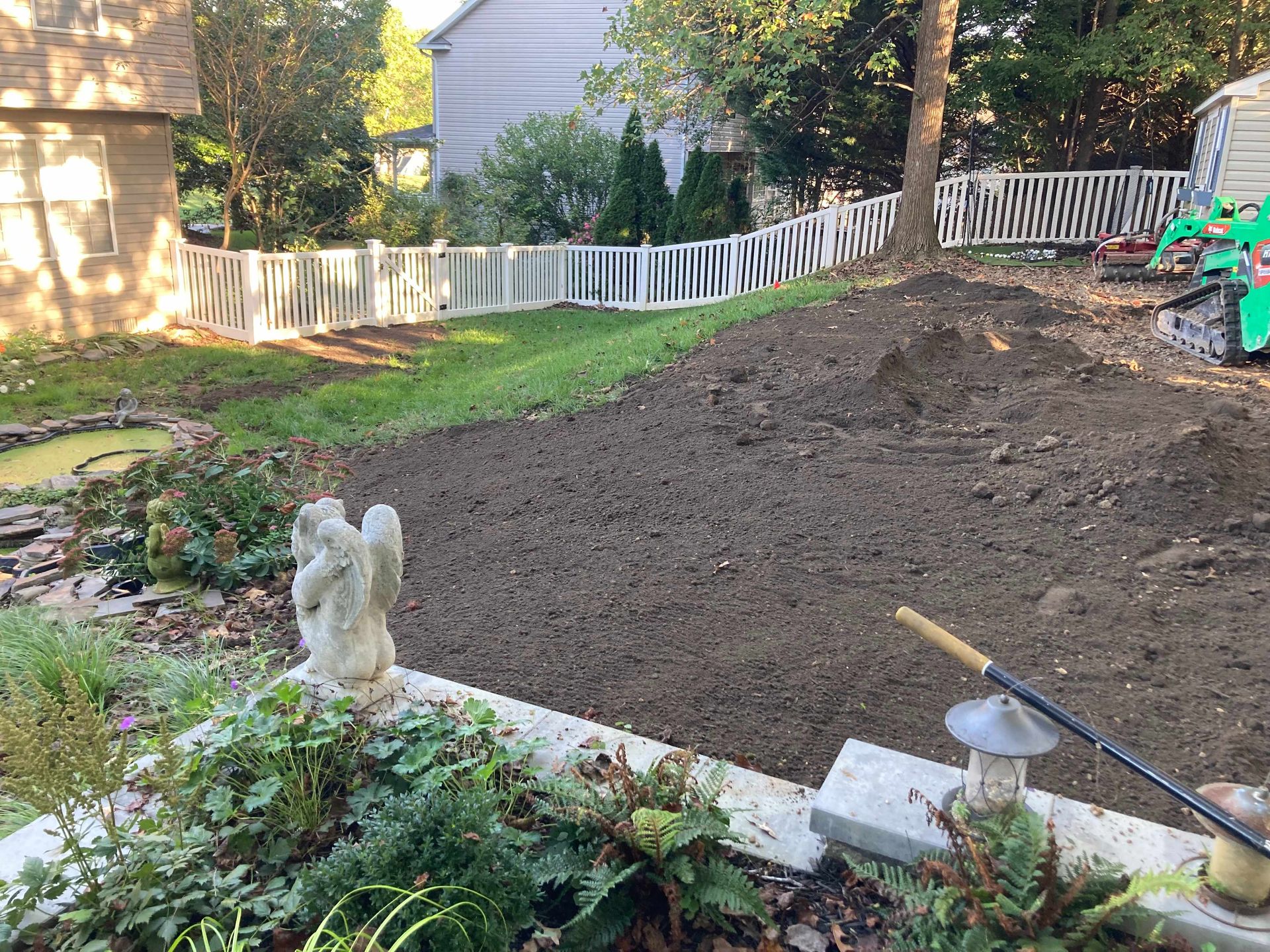 Backyard garden bed under construction with soil mound, white fence, and small statue in the foreground