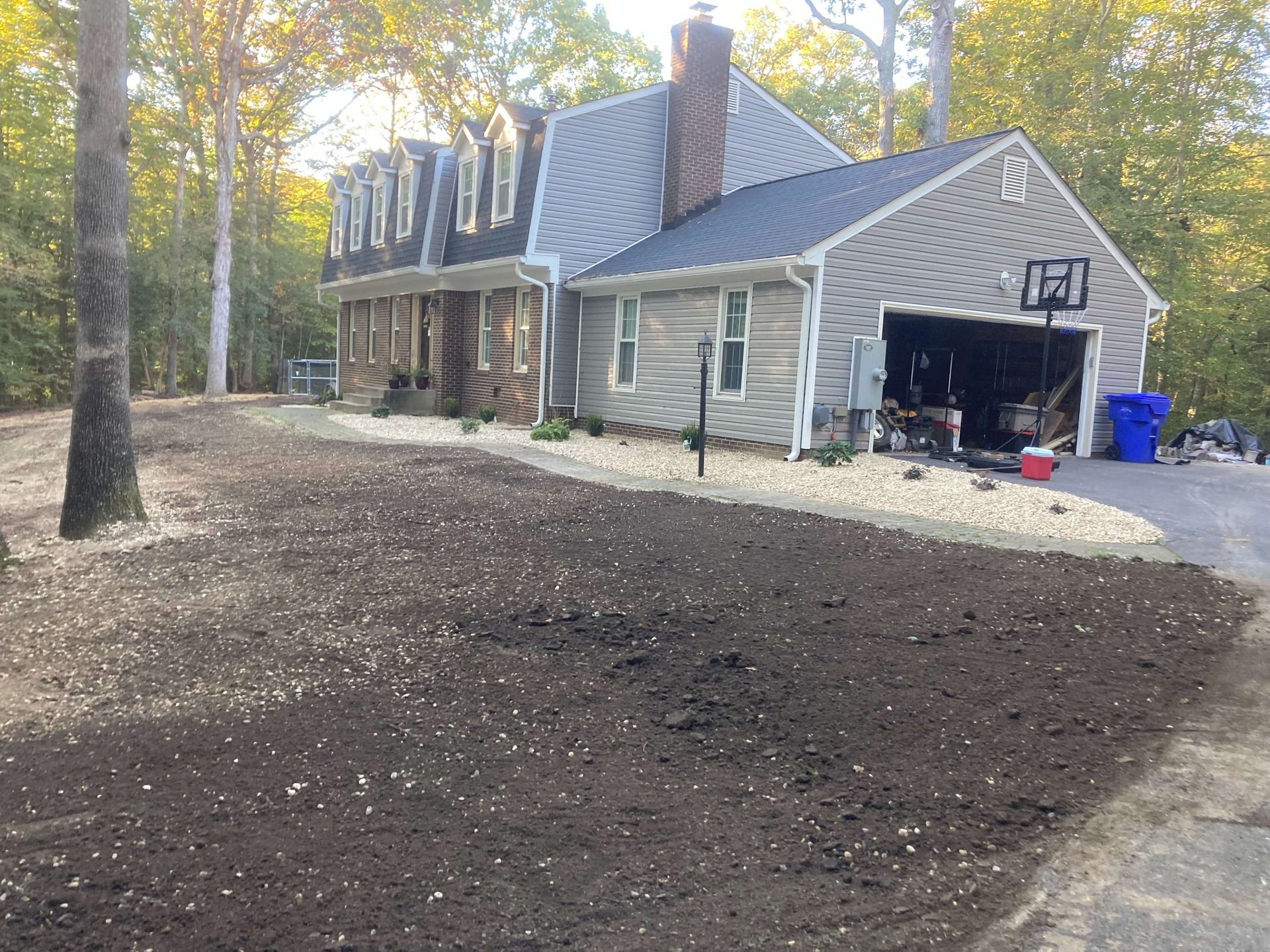 Stone house with open garage and bare dirt yard, surrounded by trees in autumn.