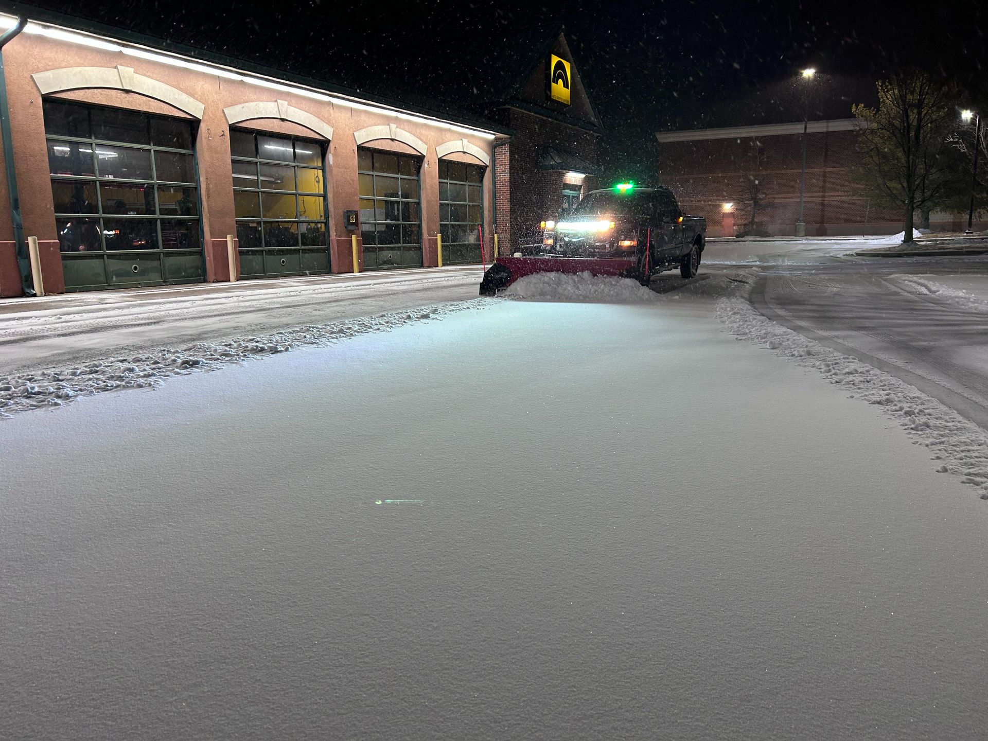 Snow-covered street at night beside lit brick buildings, with a plow truck clearing a path.