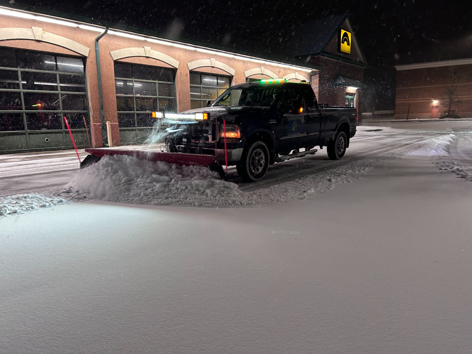 Black pickup truck plowing snow in a snowy parking lot at night beside lit storefronts