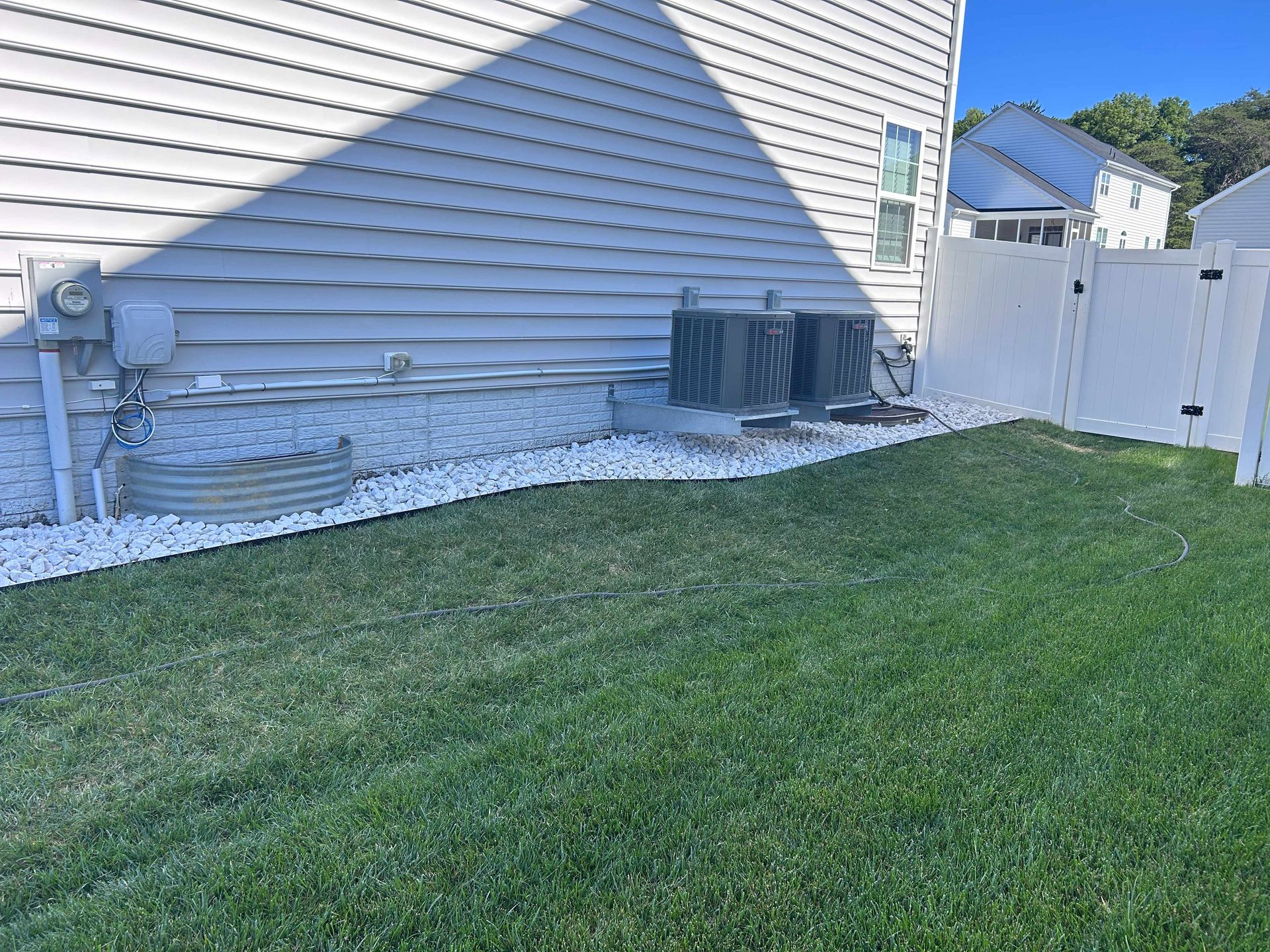Side of a house with white siding, utility units, gravel strip, grass lawn, and a white fence.