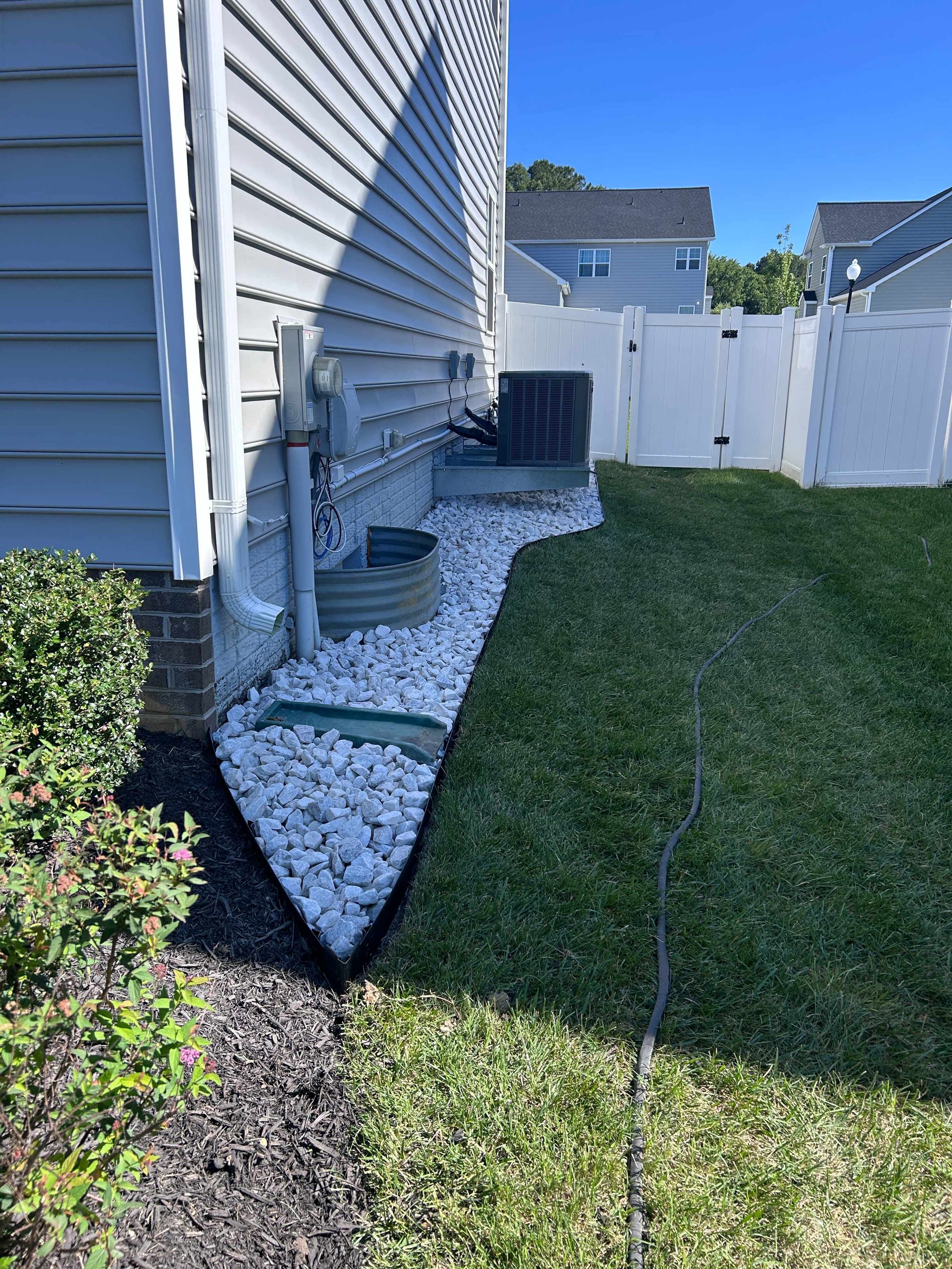 Side yard beside a house with white gravel beds, shrubs, and a green lawn under a clear blue sky