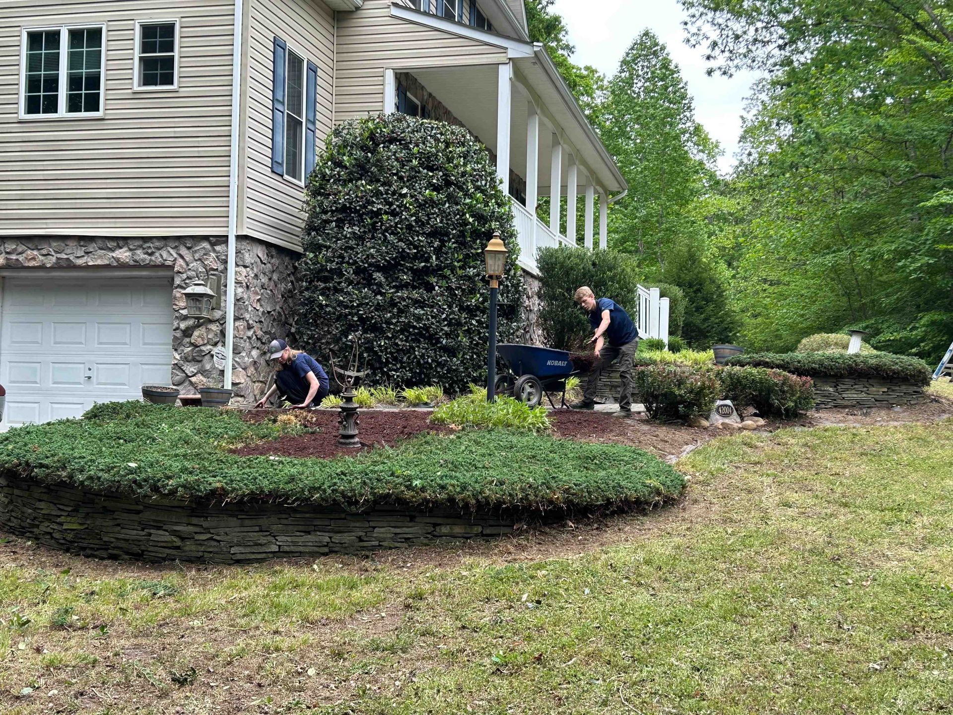 Two people tending a landscaped front yard beside a beige house with shrubs and mulch beds