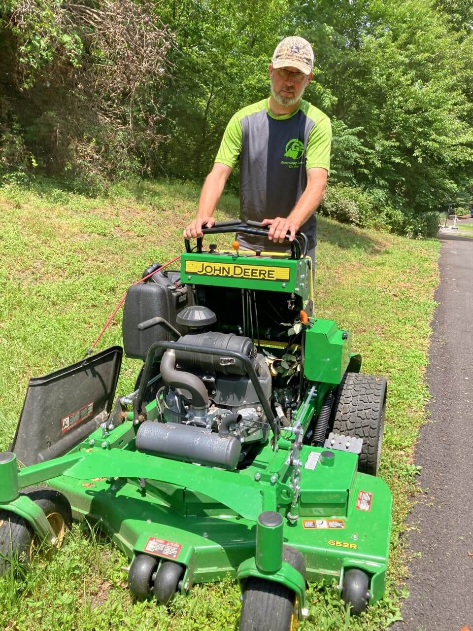 Man standing behind a green riding mower on a paved path in a wooded area