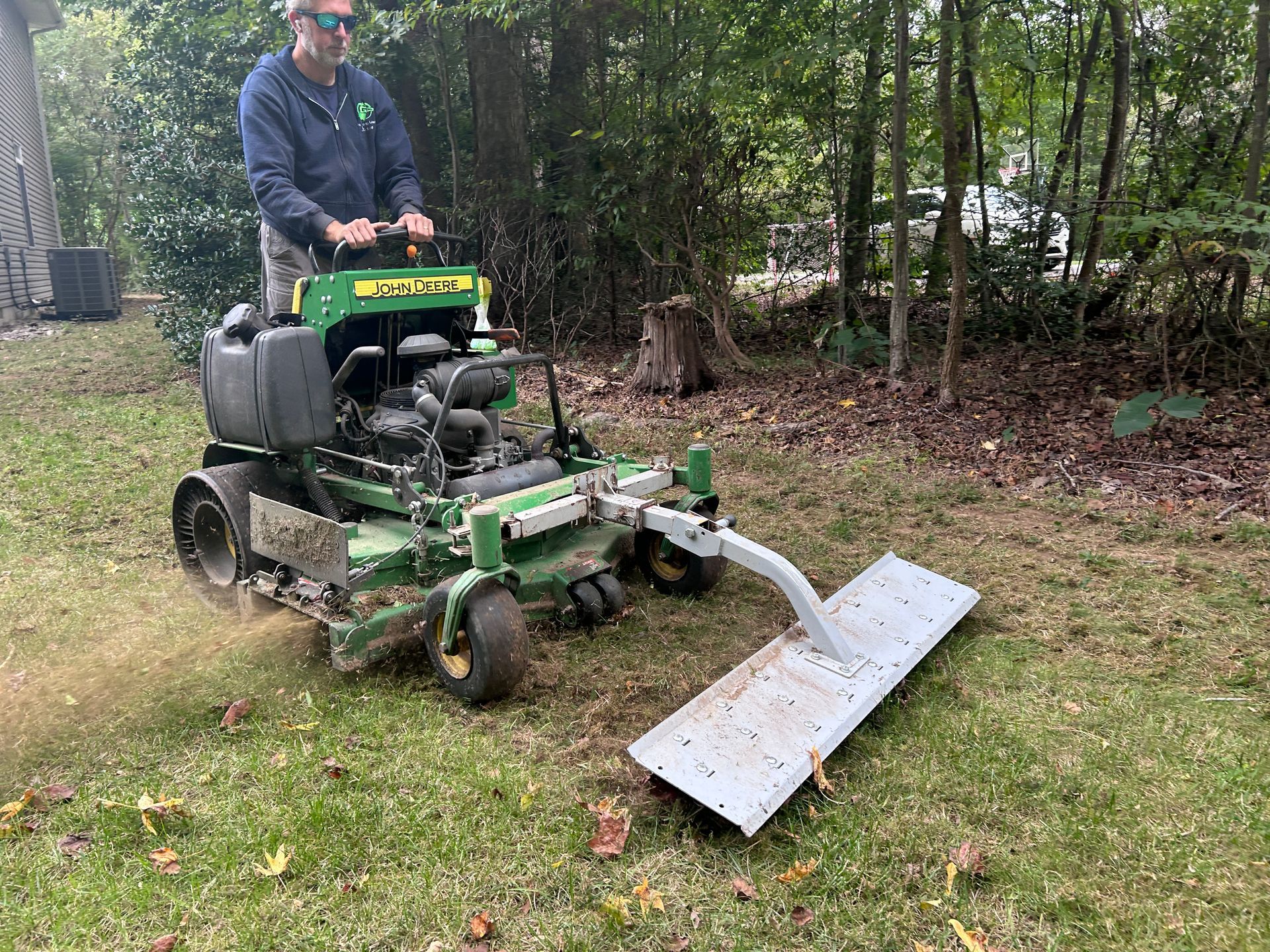 Person mowing grass with a green riding mower and front blade in a wooded yard