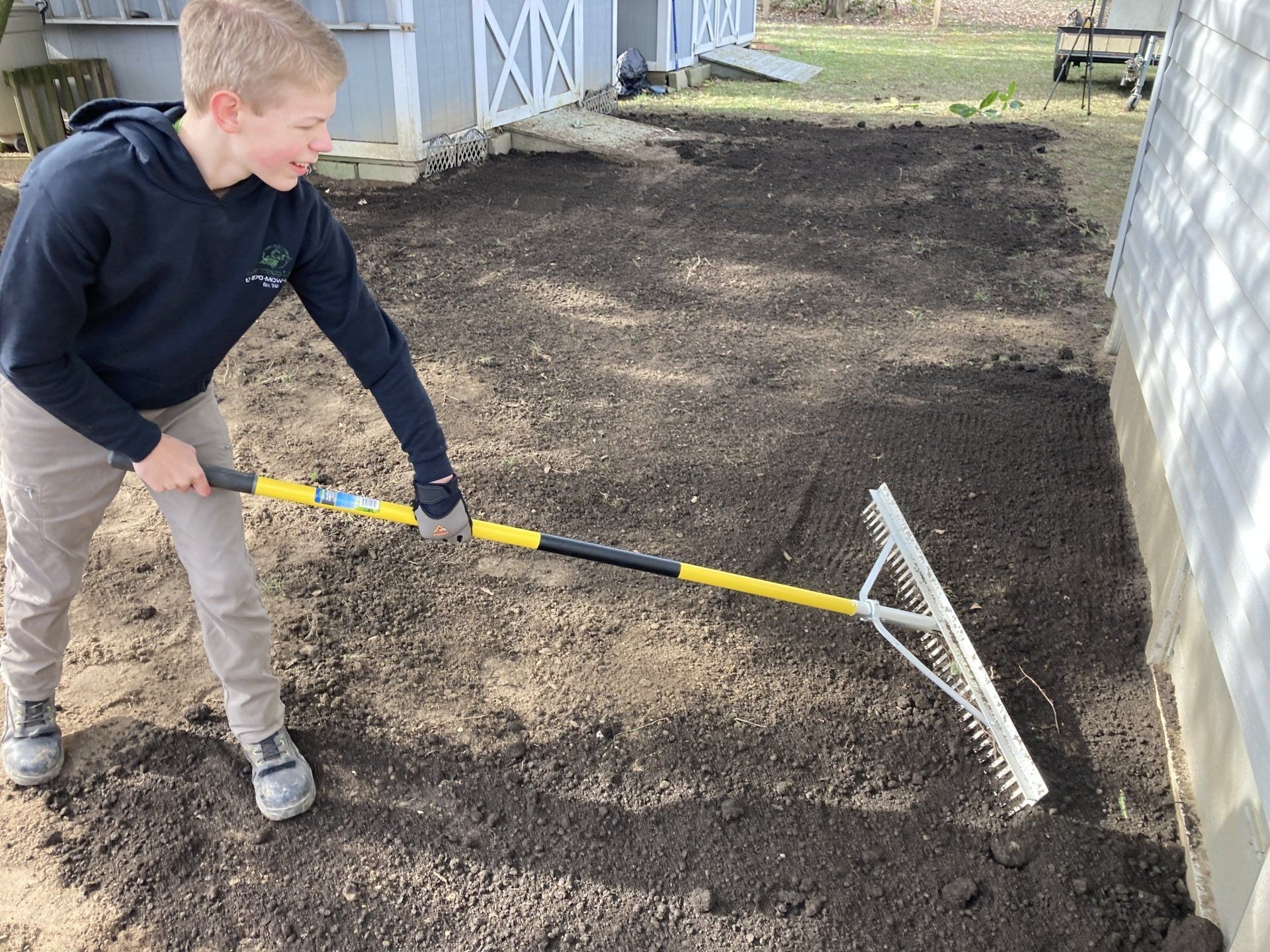 Person raking a dirt path beside a building with a wide rake