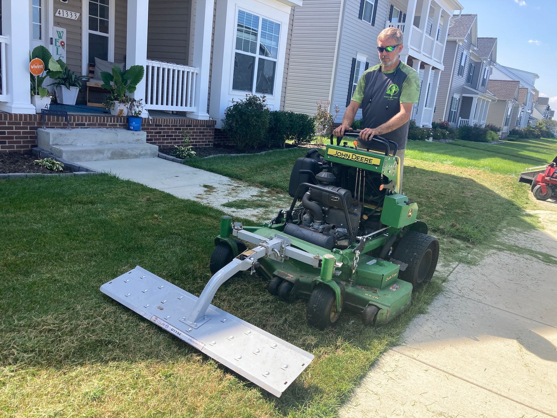 Lawn mower clearing a sidewalk edge in a suburban neighborhood, with a worker guiding it near houses.