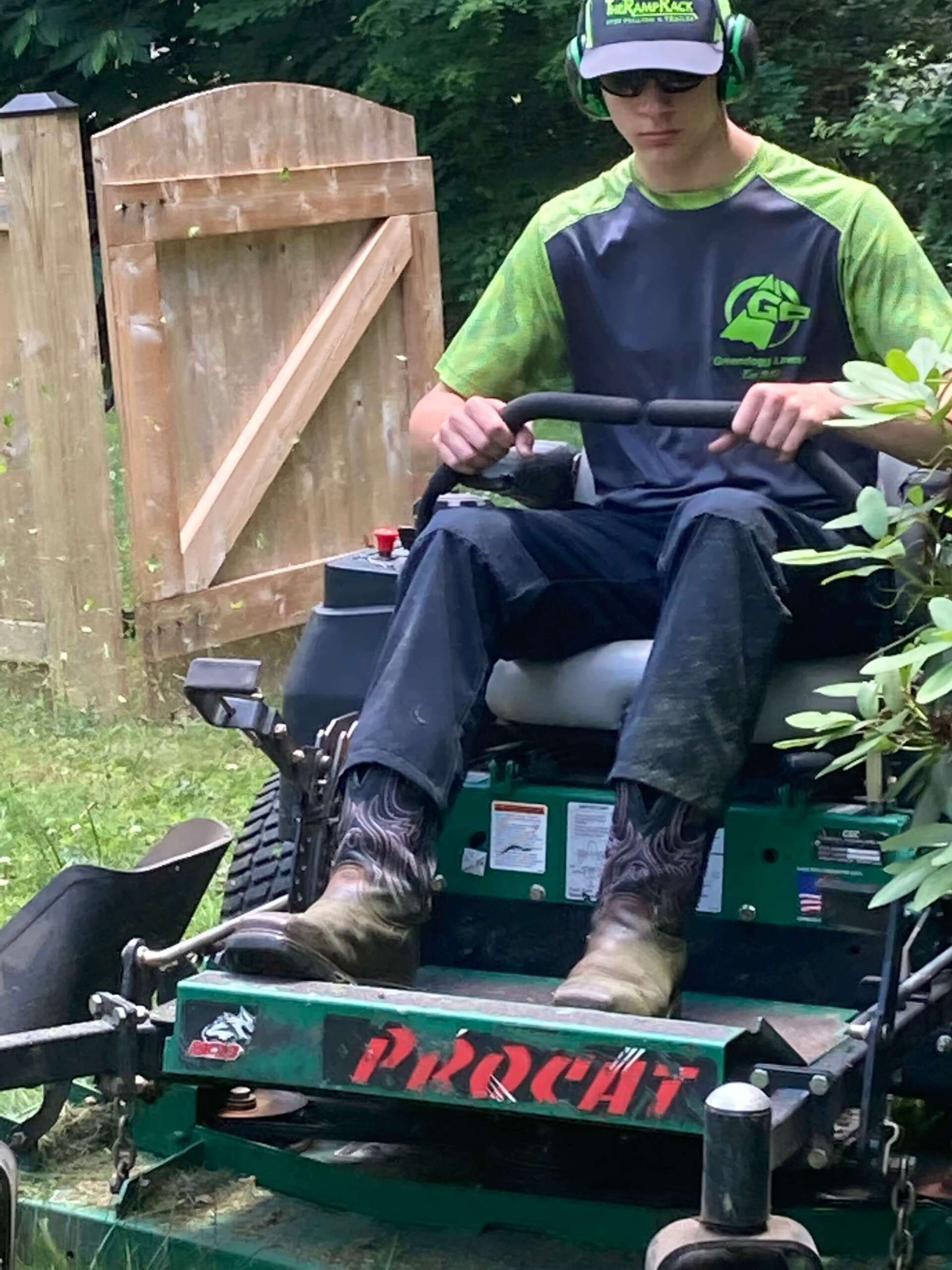 Person mowing grass on a green riding mower near a wooden gate, wearing a green shirt and cap