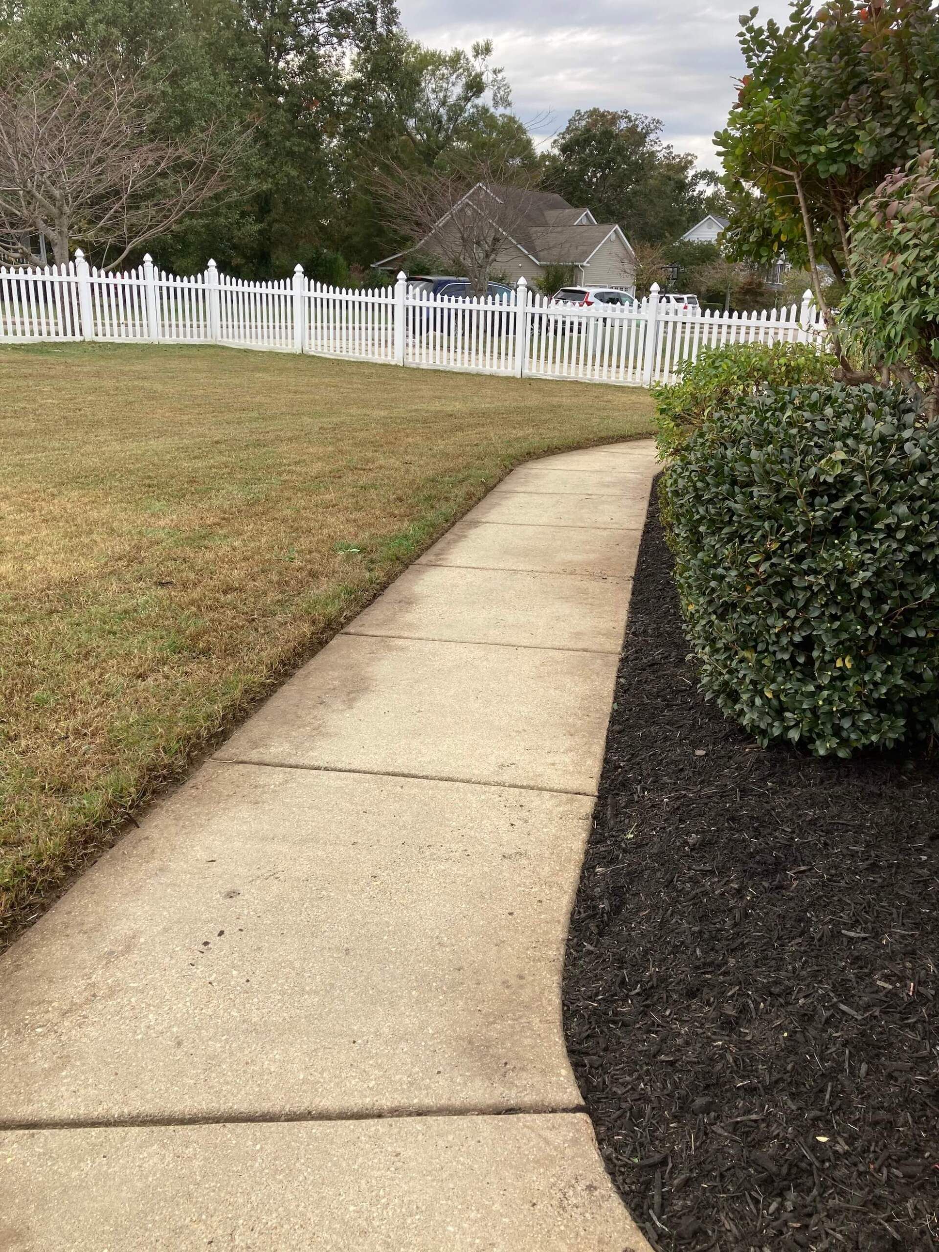 Curved concrete walkway beside a lawn, mulch bed, bushes, and white picket fence under a cloudy sky
