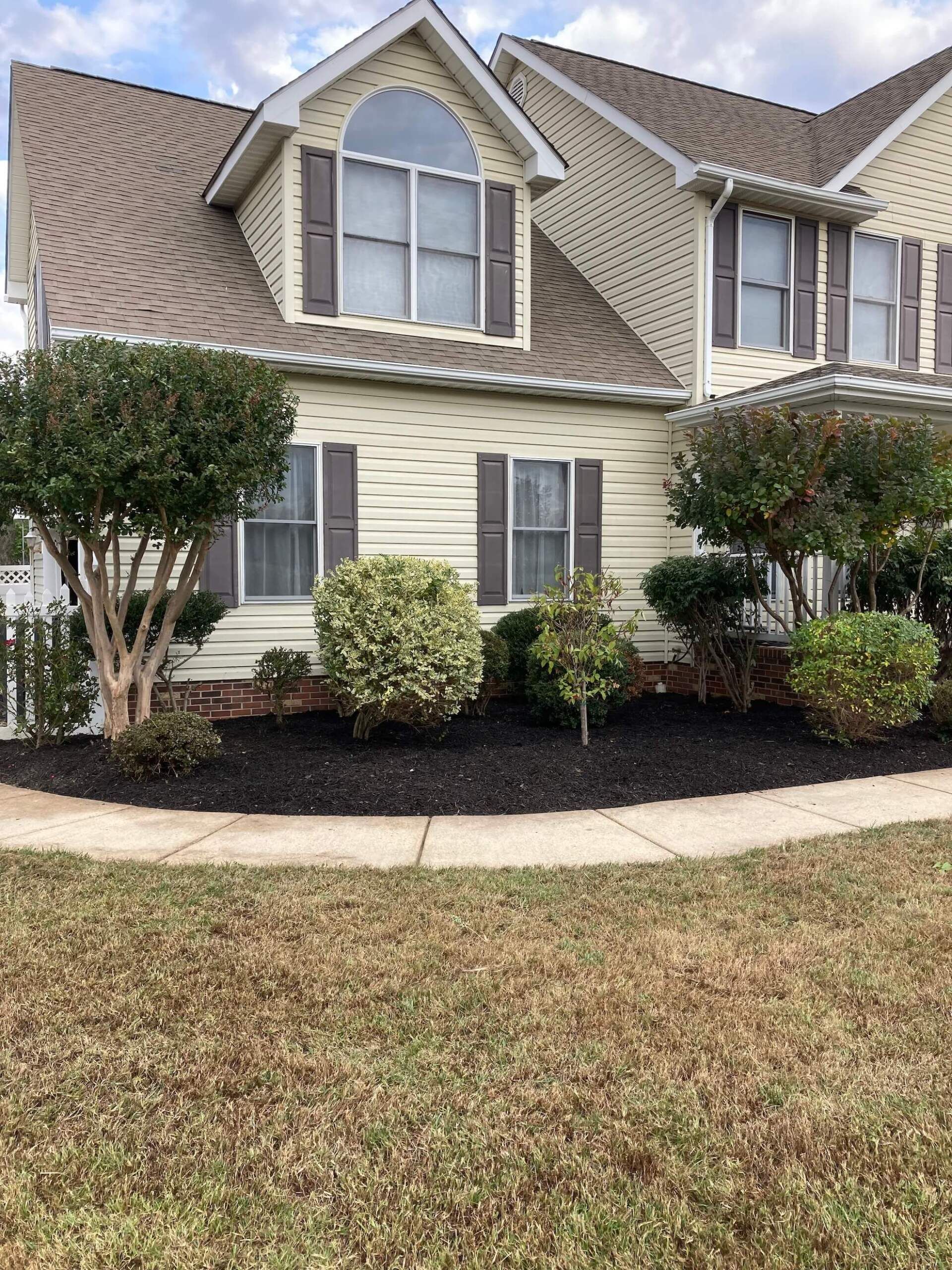 Suburban house with beige siding, steep roof, shrubs, and a curved front walkway
