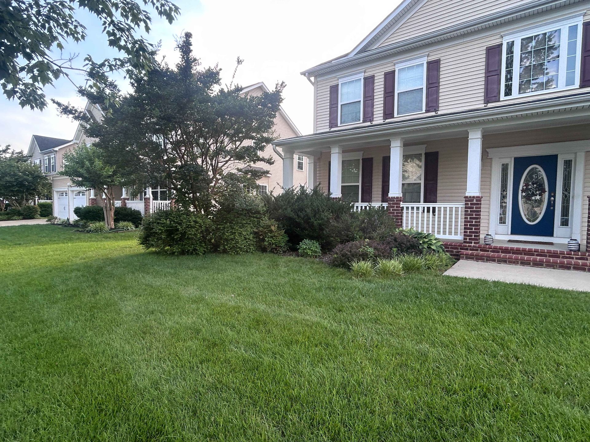 Suburban house with front porch, blue door, white columns, and a green lawn with shrubs and a tree