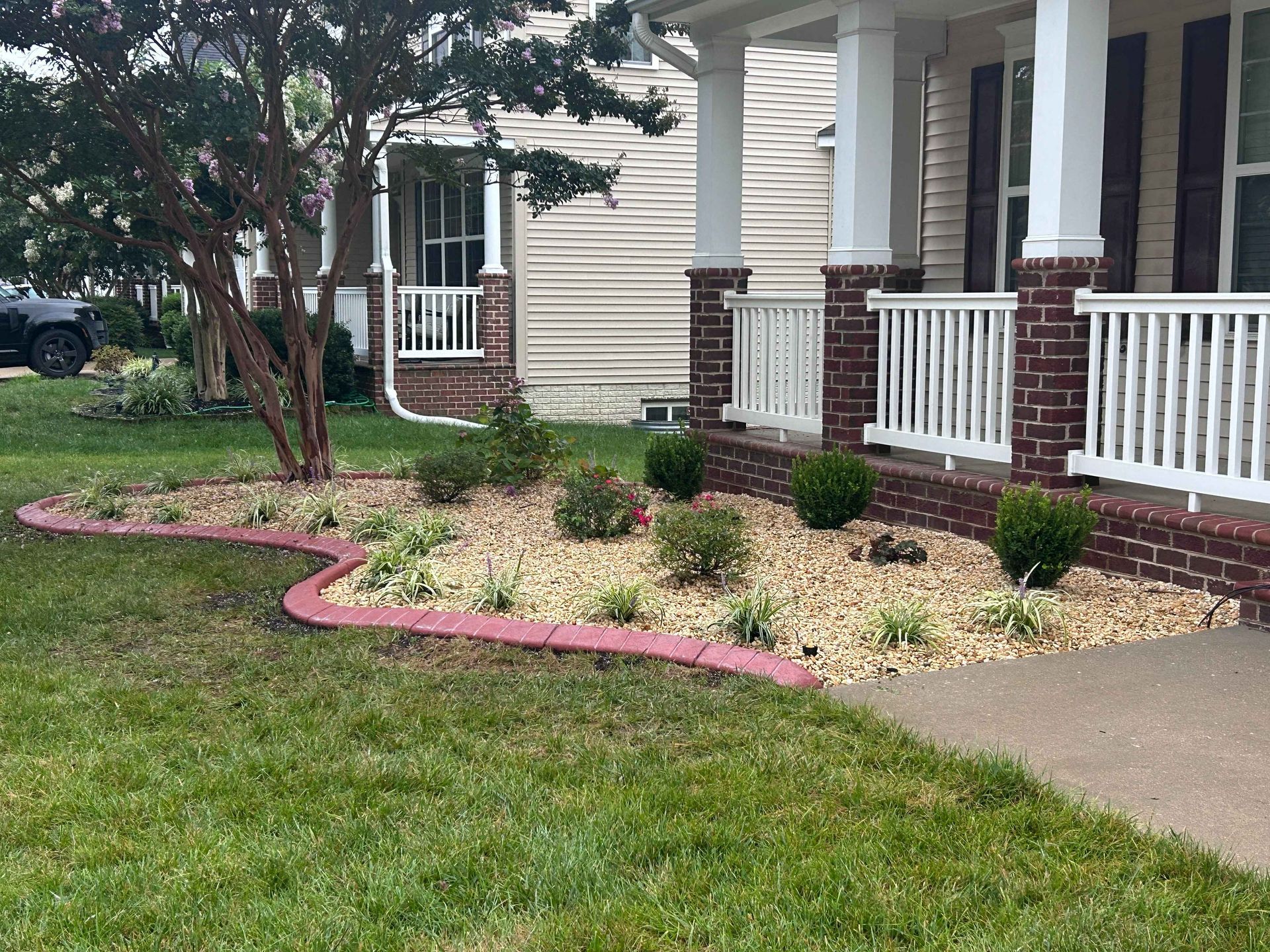 Front porch with white railing, brick columns, and a pink-edged garden bed beside a sidewalk
