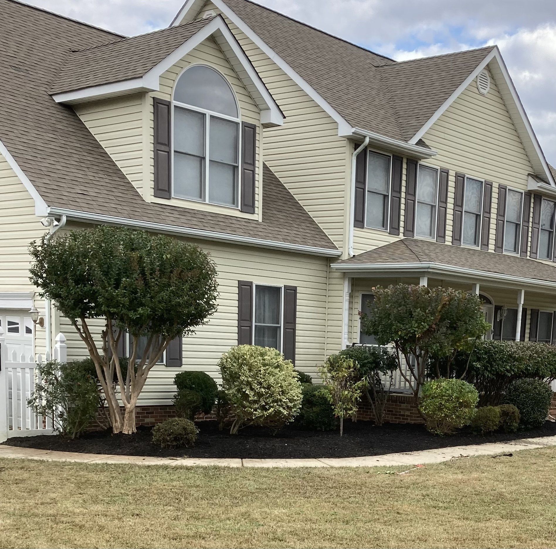 Two-story beige house with gabled roof, white trim, shutters, and trimmed shrubs in front yard