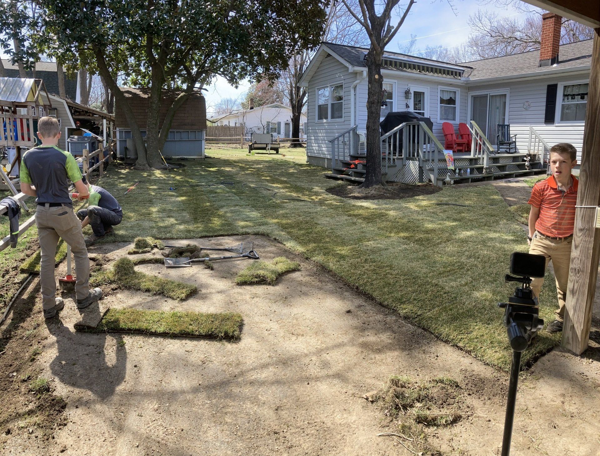 A backyard with two people standing on a dirt path beside a white house and trees on a sunny day