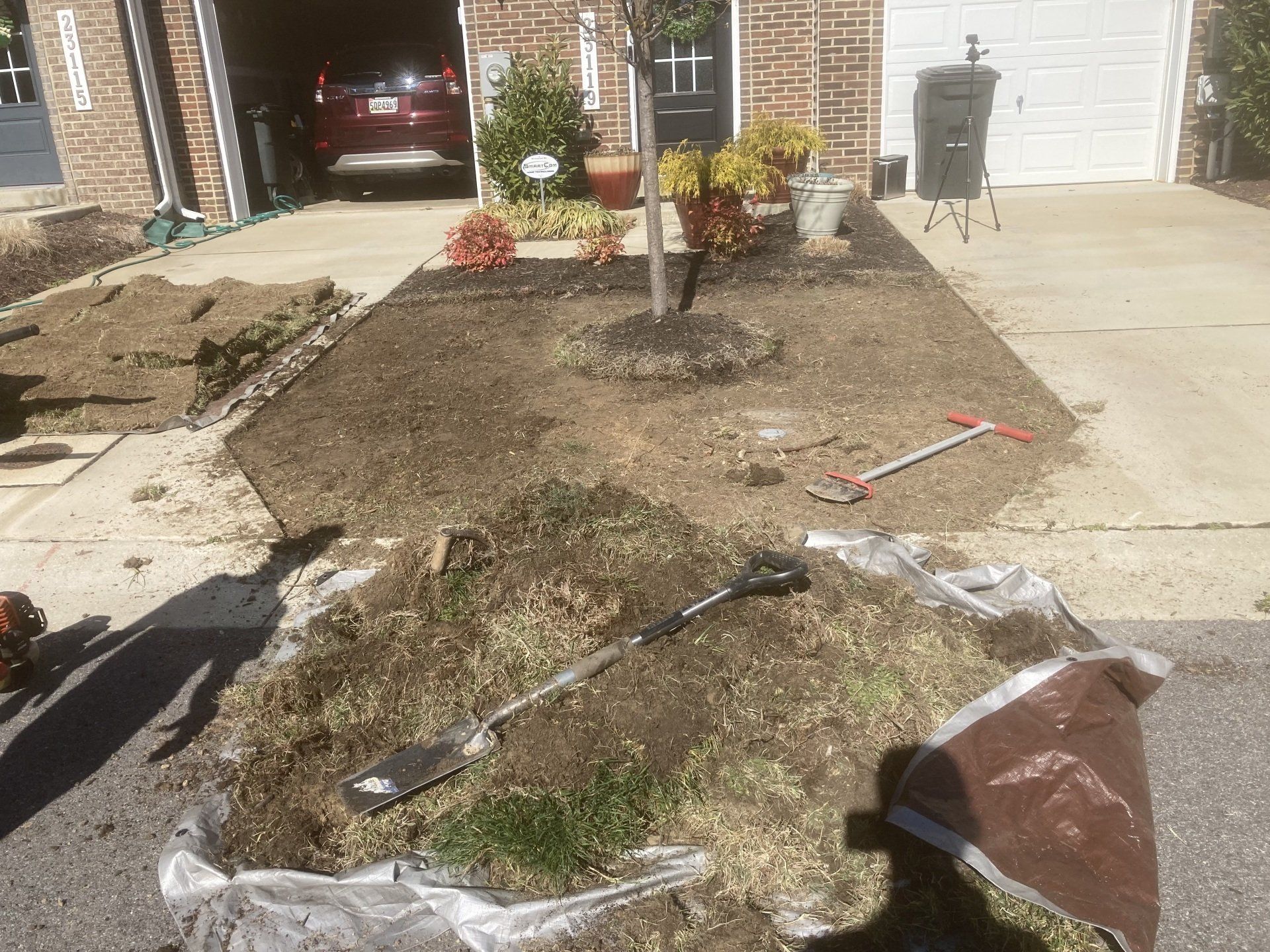Front yard with torn landscaping fabric, exposed soil, and a driveway beside a brick house.