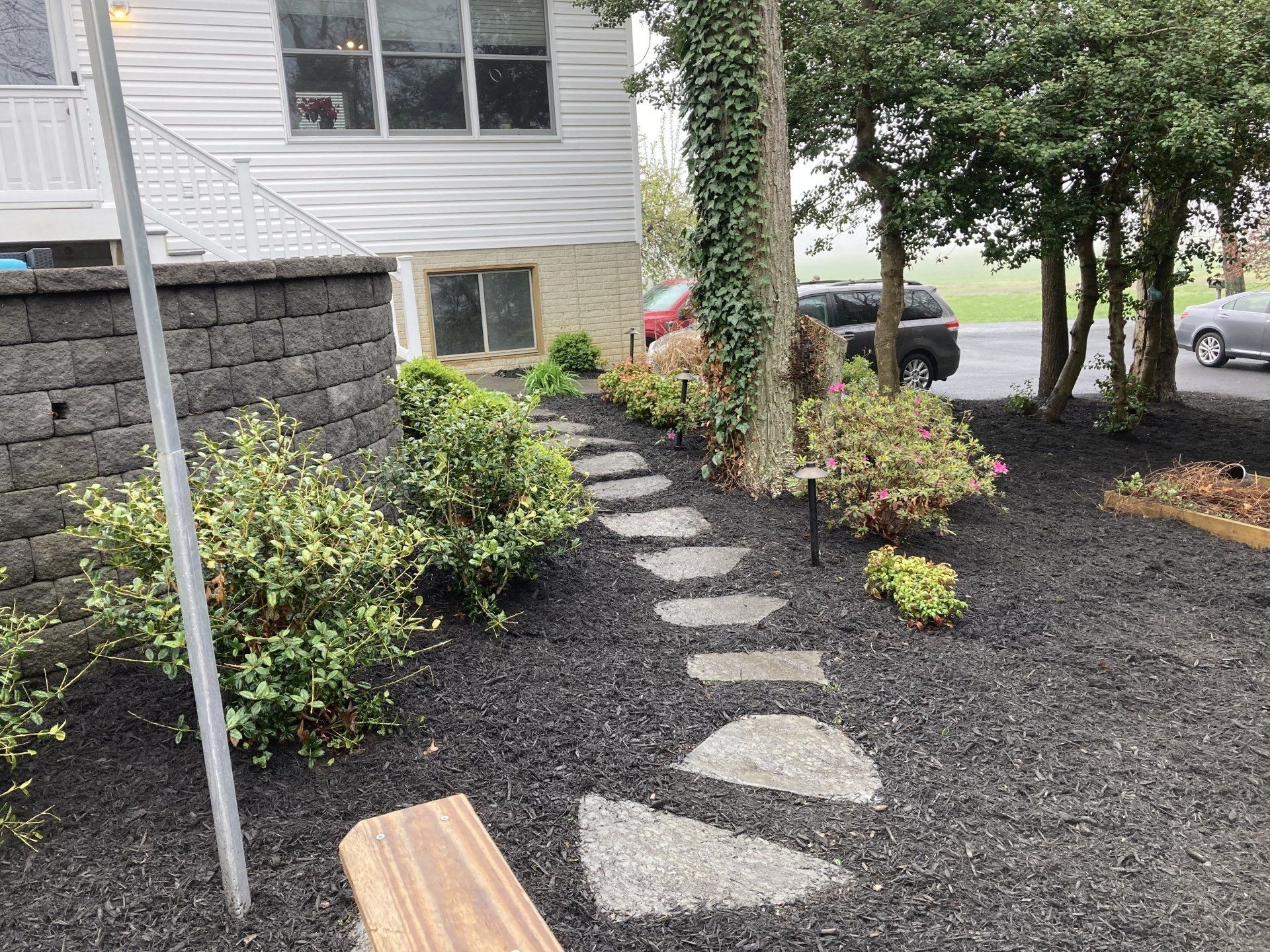 Stepping-stone path through dark mulch beside a house, with shrubs and trees in a front yard.