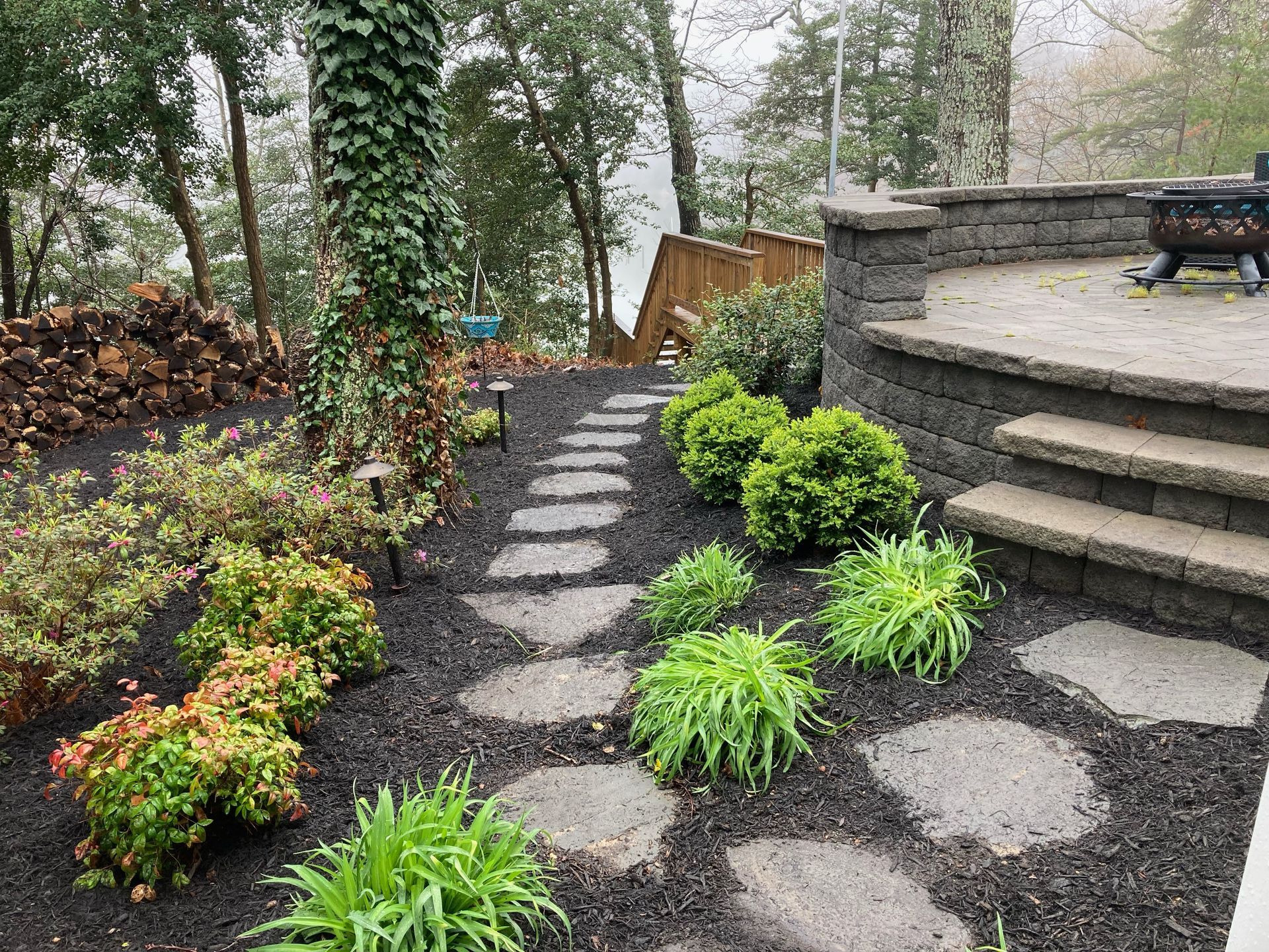 Stone stepping path through a landscaped garden with shrubs, ivy-covered trees, and a raised patio.