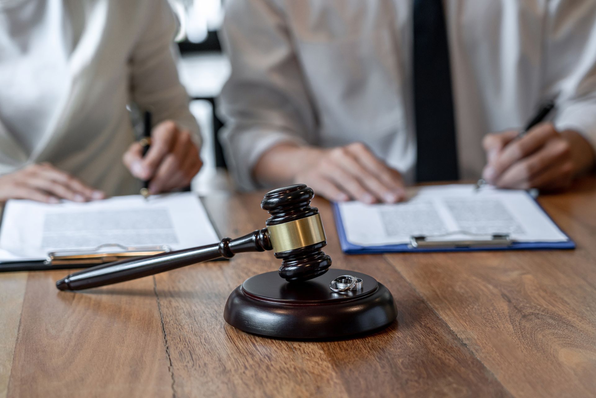 A pair of spouses sit in front of a table with a gavel on top of it, with two wedding rings. Judge’s gavel on a wooden desk with two people signing documents in the background