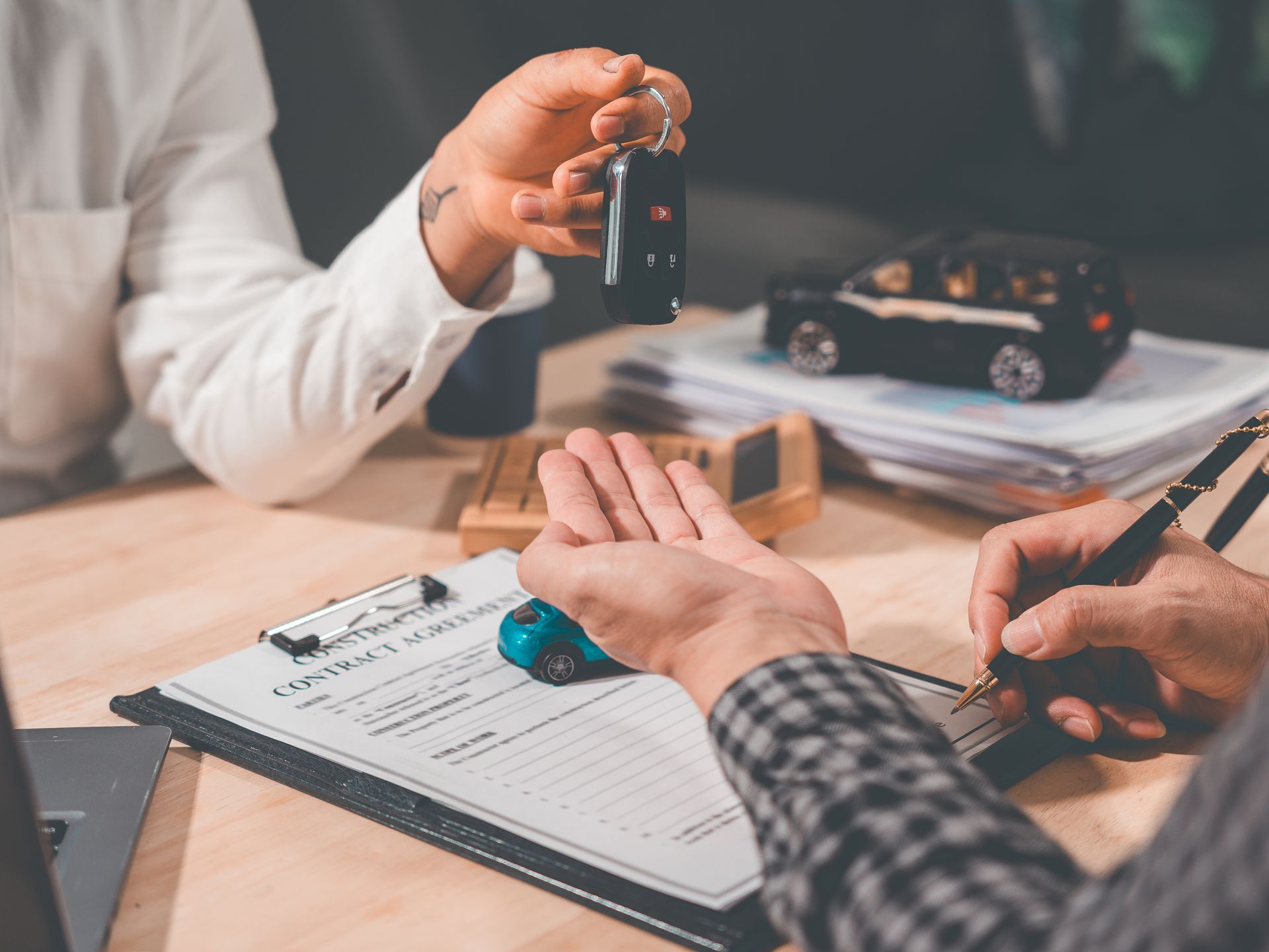 Hands signing a car sale form while a dealer holds out keys over a desk with a model car. Hands signing a car sale form while a dealer holds out keys over a desk with a model car.