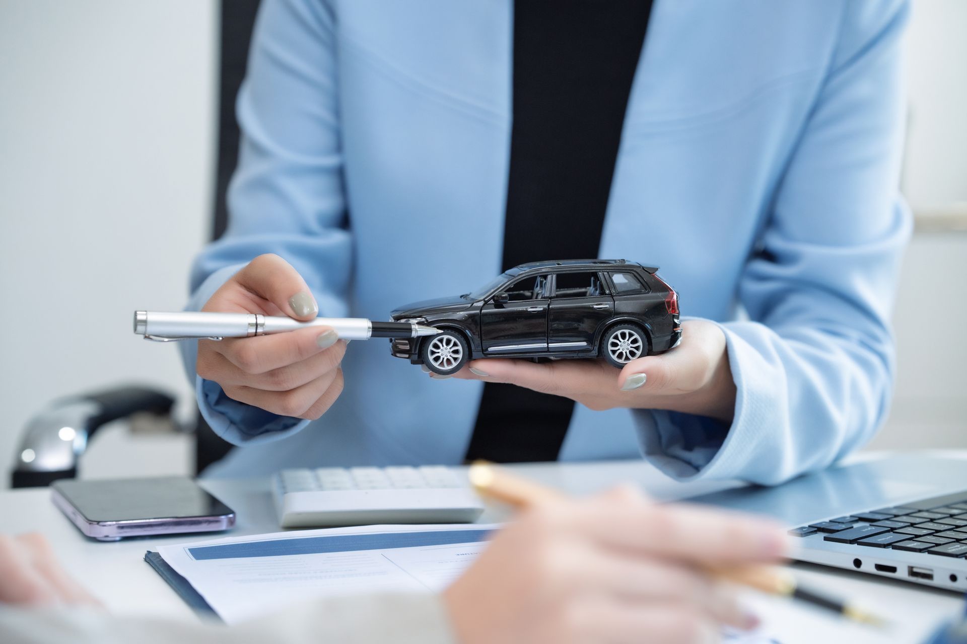Person in a blue blazer presenting a black car model with a pen at a desk in an office setting Person in a blue blazer presenting a black car model with a pen at a desk in an office setting