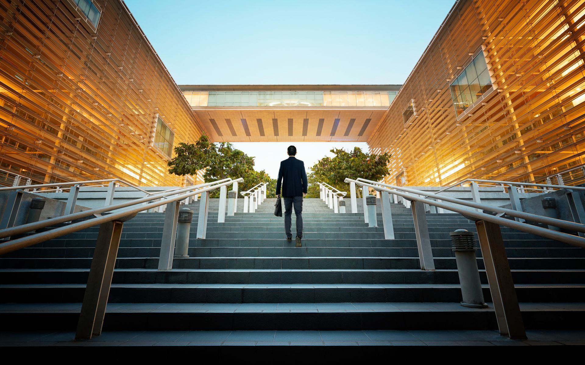 A lone figure in a suit walks up wide, stone steps toward a bridge connecting two modern buildings with glowing slats. A lone figure in a suit walks up wide, stone steps toward a bridge connecting two modern buildings with glowing slats.