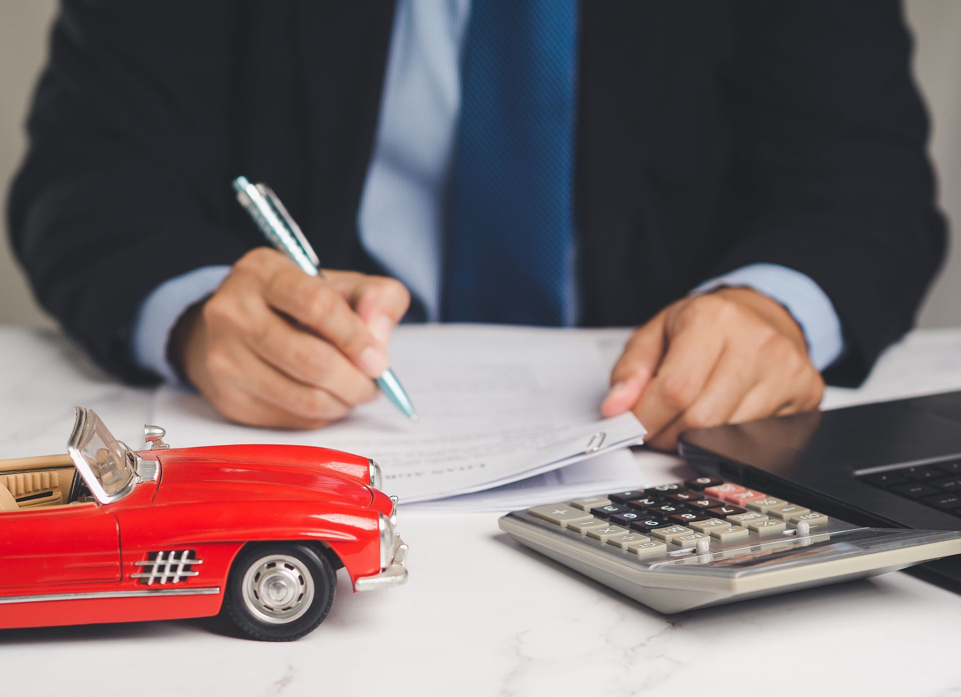 Businessperson writing on documents beside a calculator, laptop, and red toy car Businessperson writing on documents beside a calculator, laptop, and red toy car