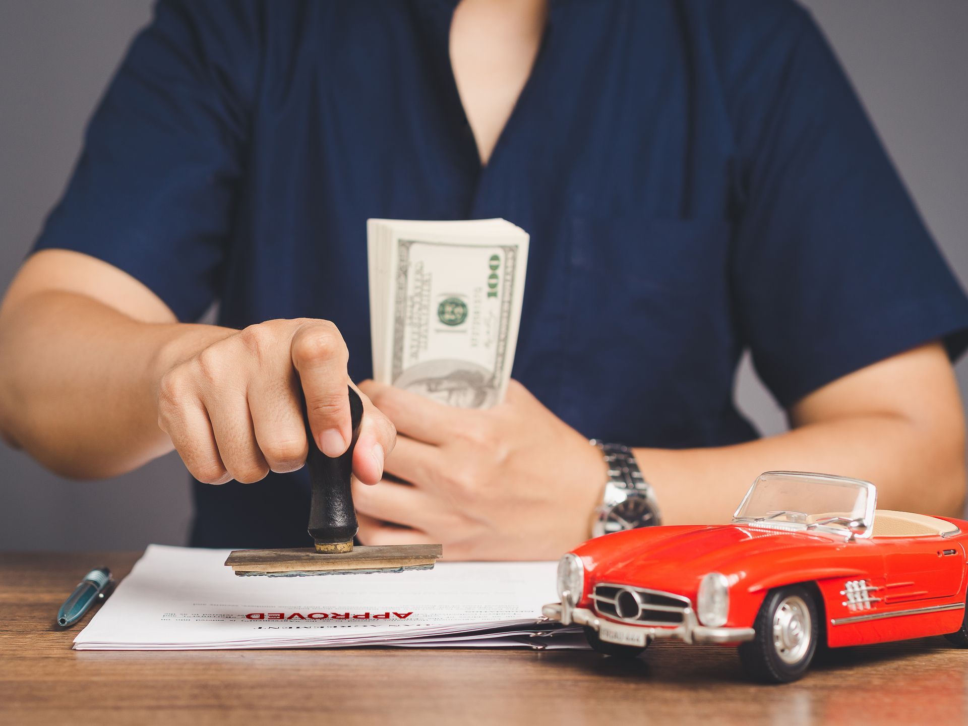 A person stamping a document near a stack of cash and a red toy car on a desk. A person stamping a document near a stack of cash and a red toy car on a desk.