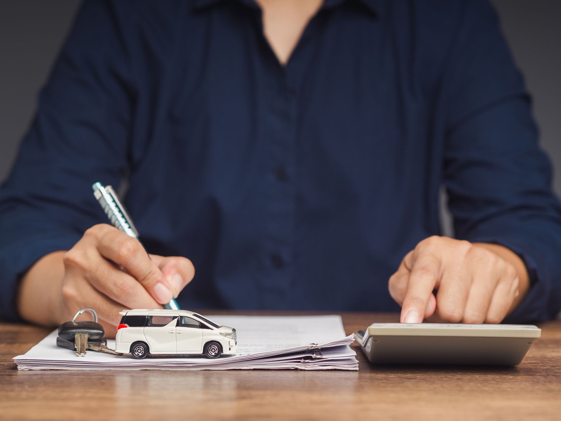 Person writing on paperwork at a desk beside a toy car and calculator