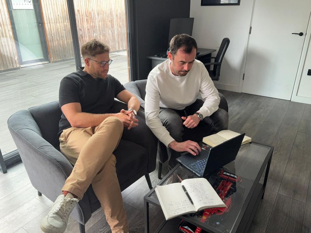 Two people sit on a sofa reviewing a laptop and open notebook in a bright room.