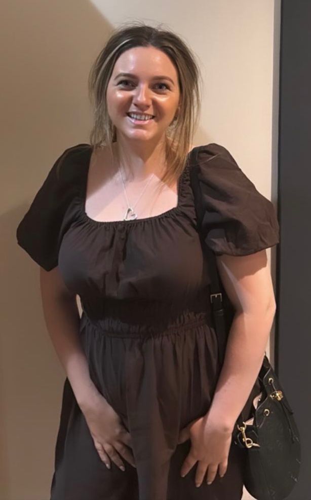 Smiling woman in a black dress standing against a beige wall, holding a black handbag.