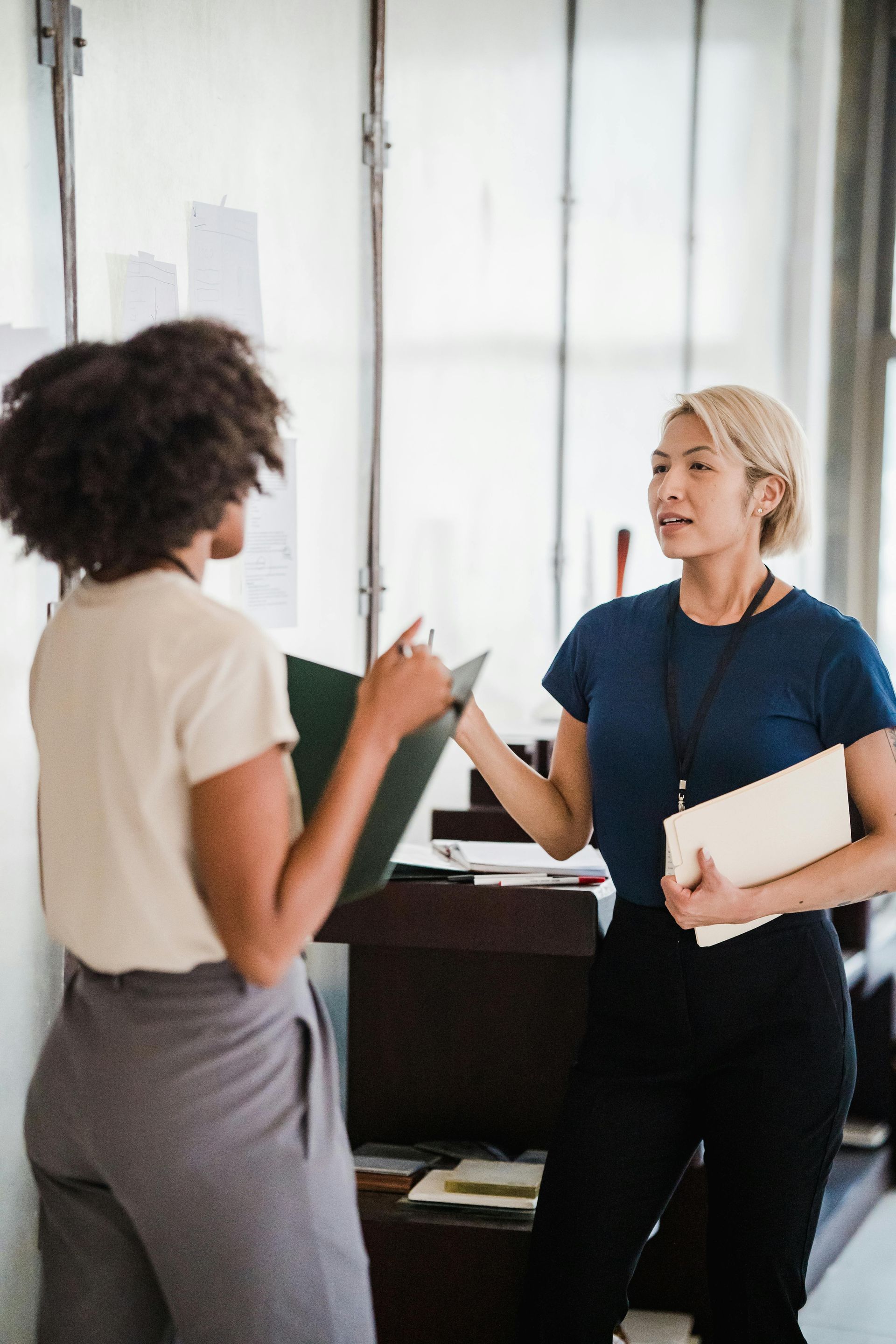 Two people talking in an office, one holding a notebook and the other gesturing with papers.