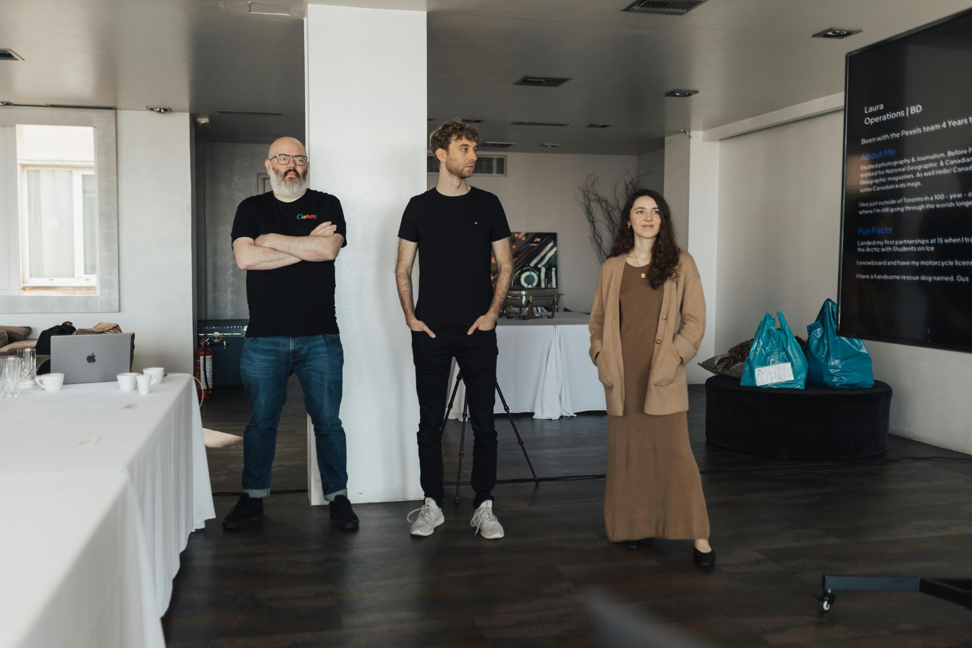 Three people standing in a modern room near a projector screen, one with folded arms.