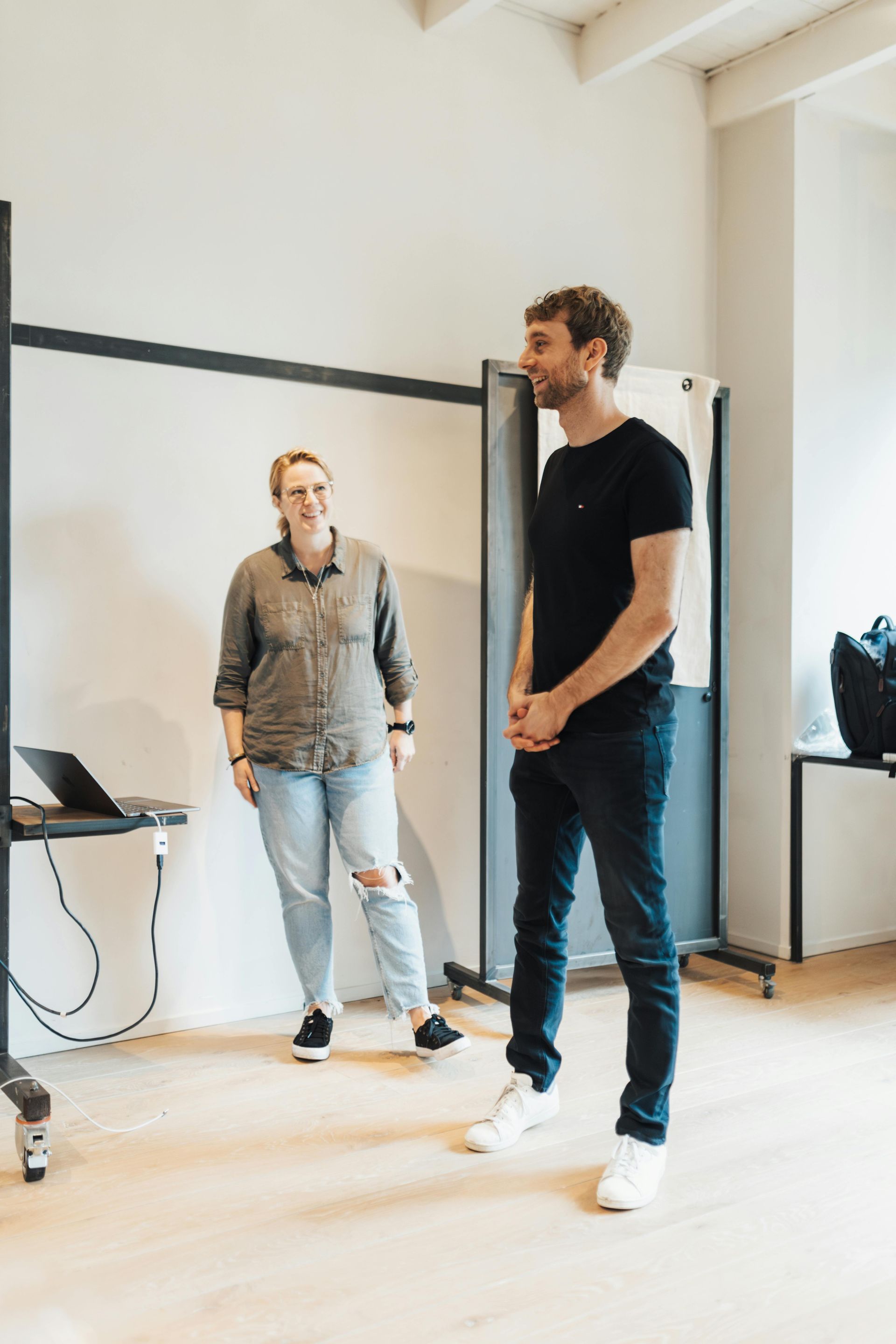 Two people standing in a bright room near a large wall mirror, with a laptop on a small table.