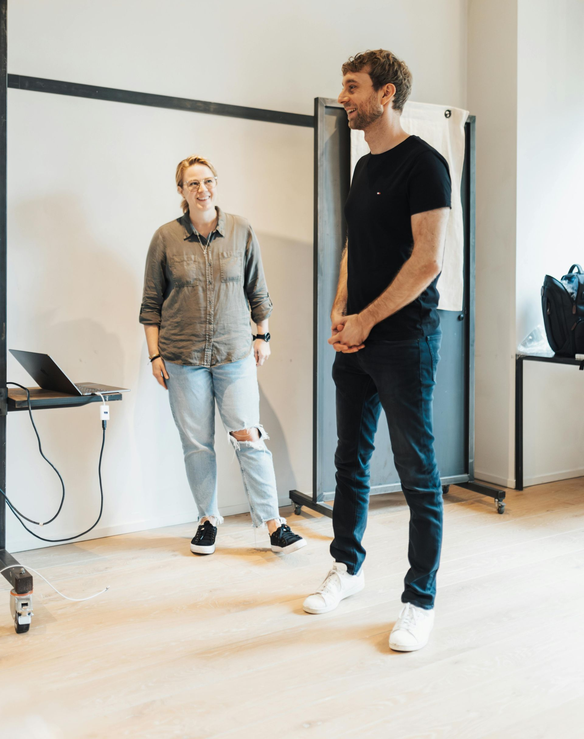 Two people standing in a bright room beside a whiteboard, one near a laptop and cables.