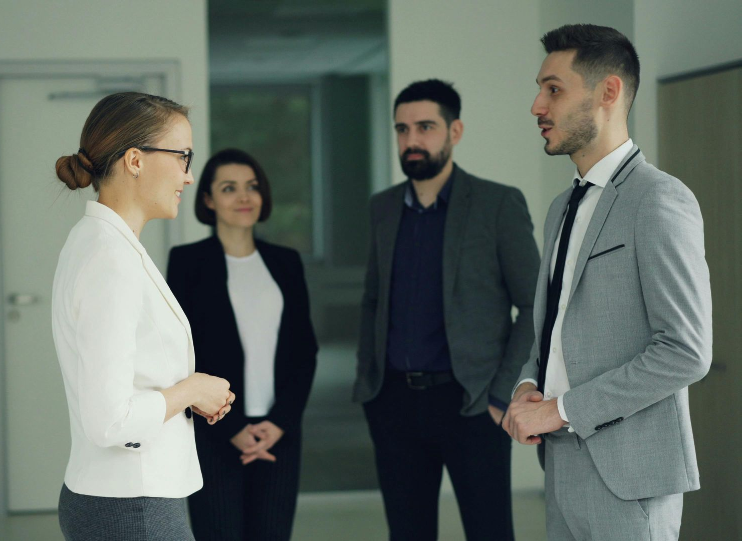 Four professionals in a meeting, two speaking in a bright office hallway.