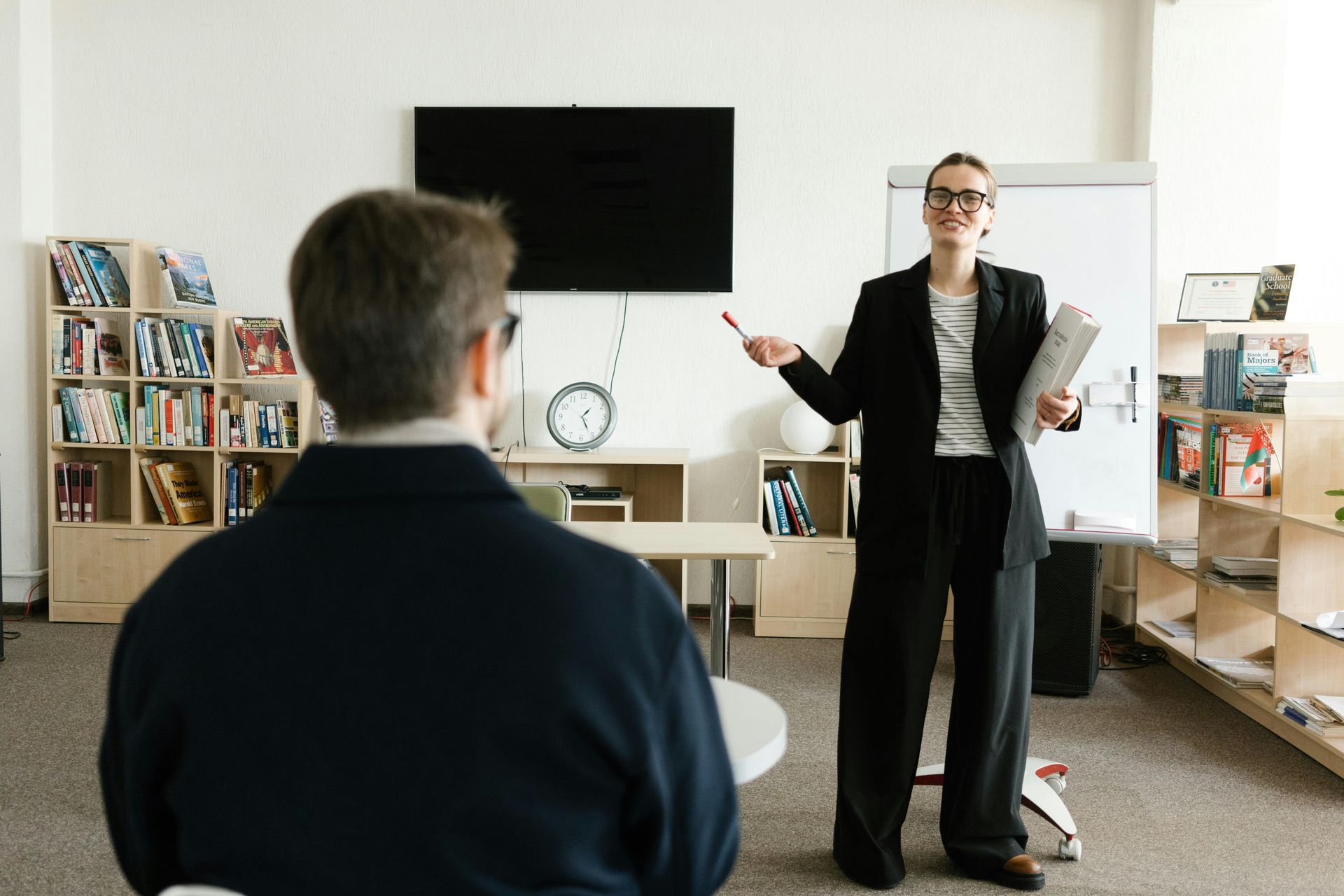 Presenter speaking in a meeting room, facing an audience seated in front of a TV and shelves.