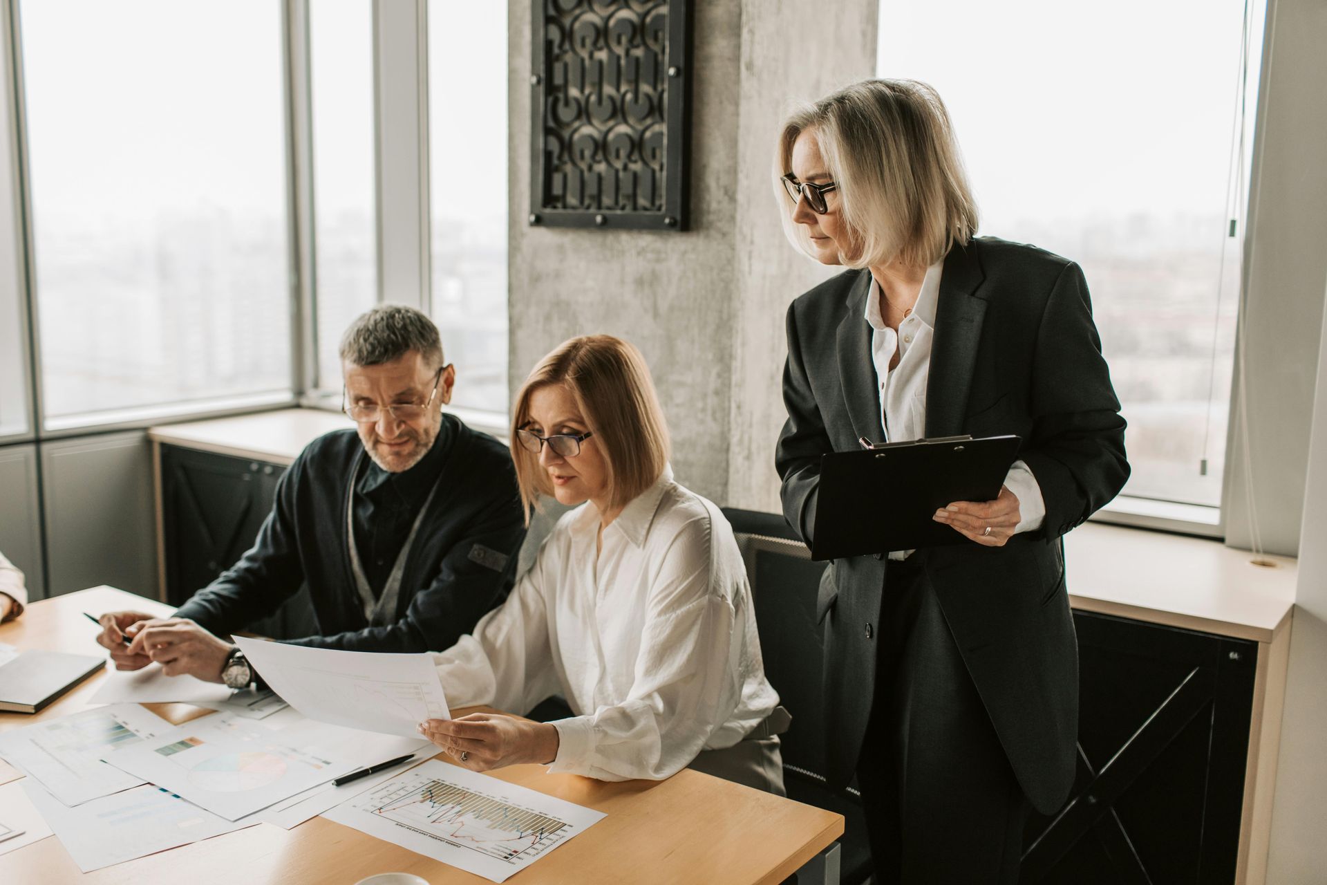Three coworkers in a bright office reviewing documents at a table, one standing with a clipboard.