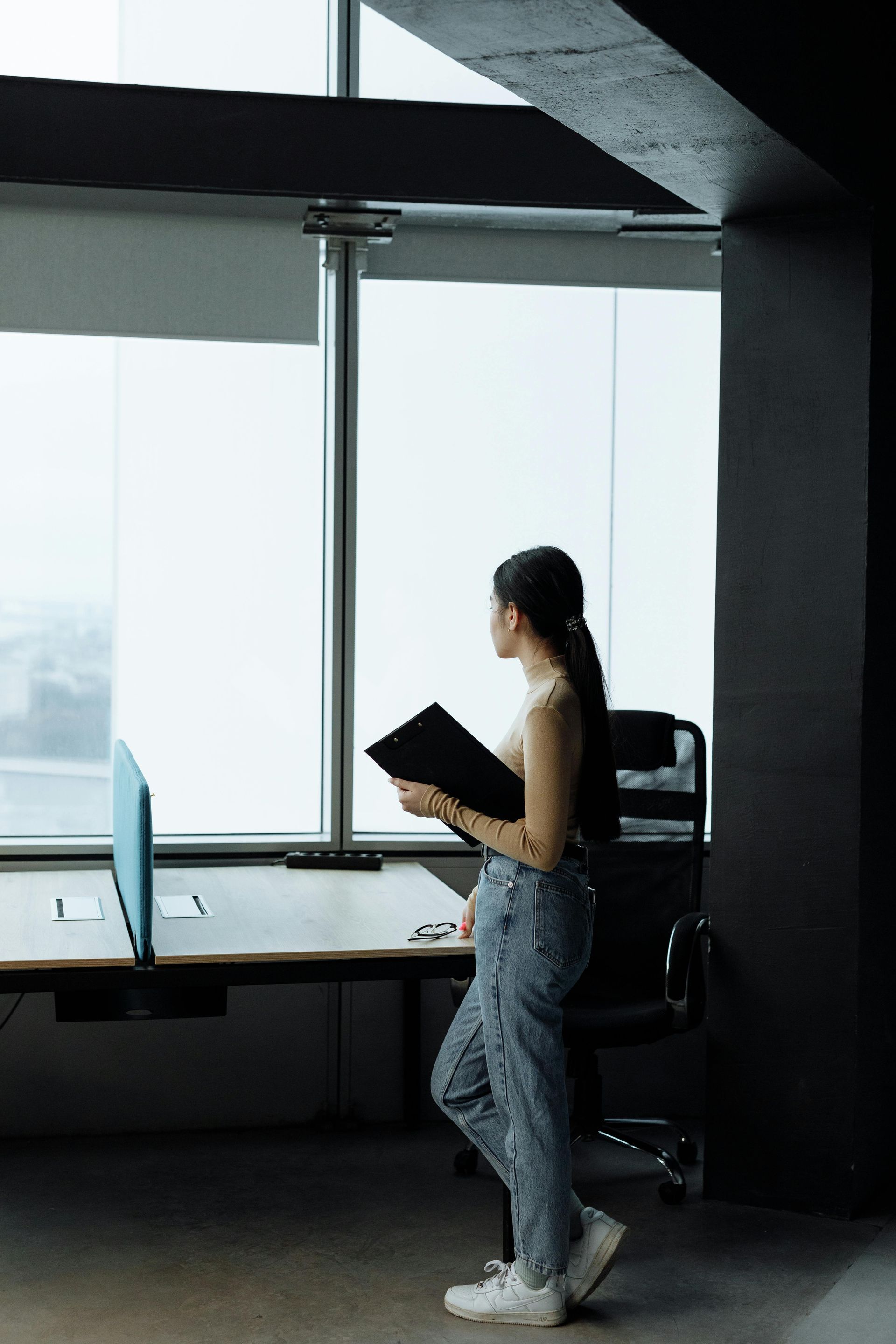 Person standing by a large window, reading a black folder in a modern office.