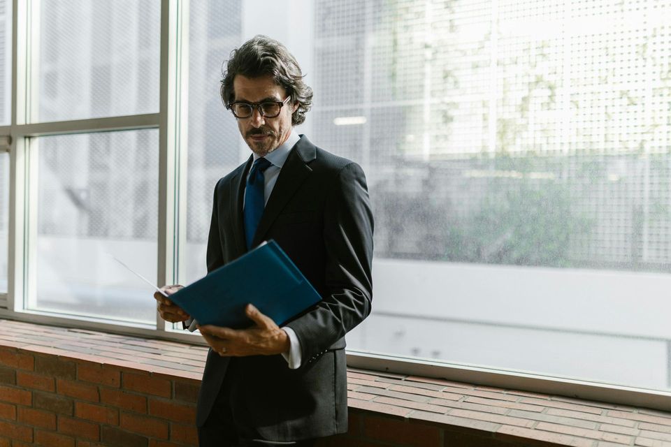 Man in a suit holding a blue folder by a bright office window